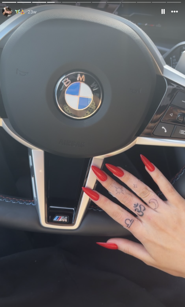 Harper Lockwood showcases her collection of delicate finger tattoos—including a sacred Om symbol, celestial motifs, and a Libra zodiac sign—resting her hand with pointed red stiletto nails on a BMW steering wheel.