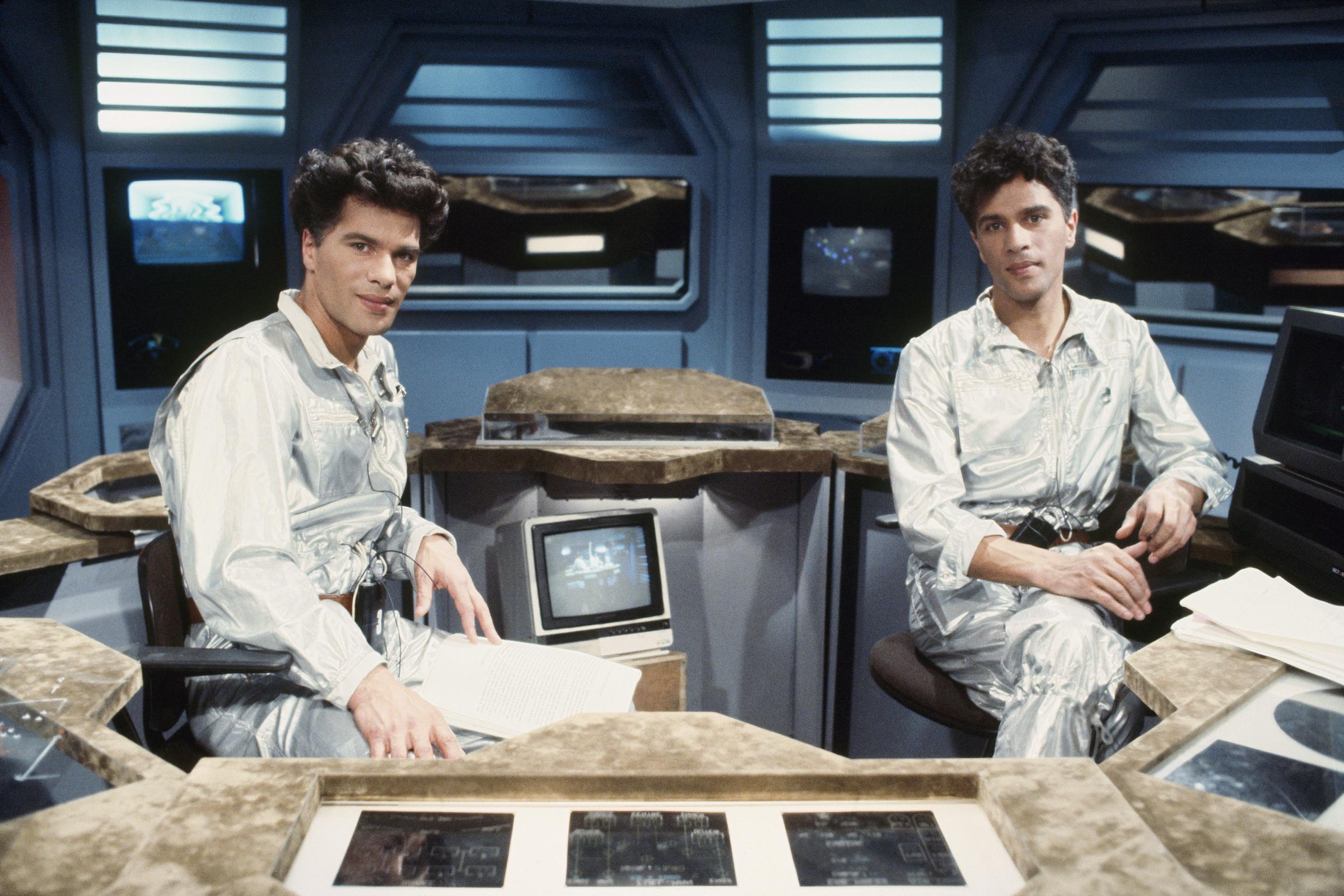 Igor and Grichka Bogdanoff on the set of their 1970s sci-fi show "Temps X," where they first rose to fame. The twins appear youthful, with chiseled cheekbones and thick, tousled hair as they pose in the studio.