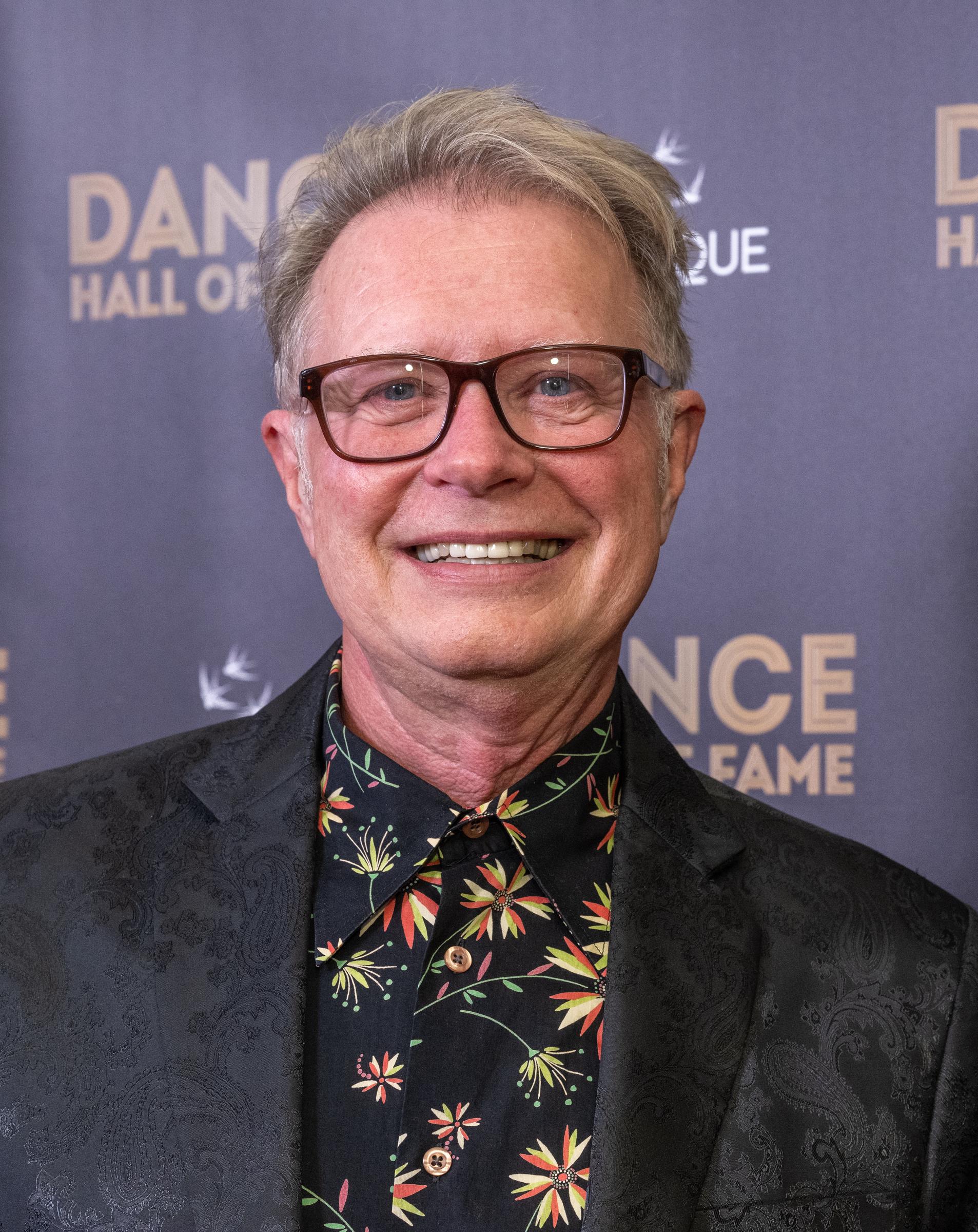 Vincent Paterson smiles for a portrait at the Dance Hall of Fame ceremony, wearing dark-framed glasses, a black patterned blazer, and a floral-print shirt. | Source: Getty Images