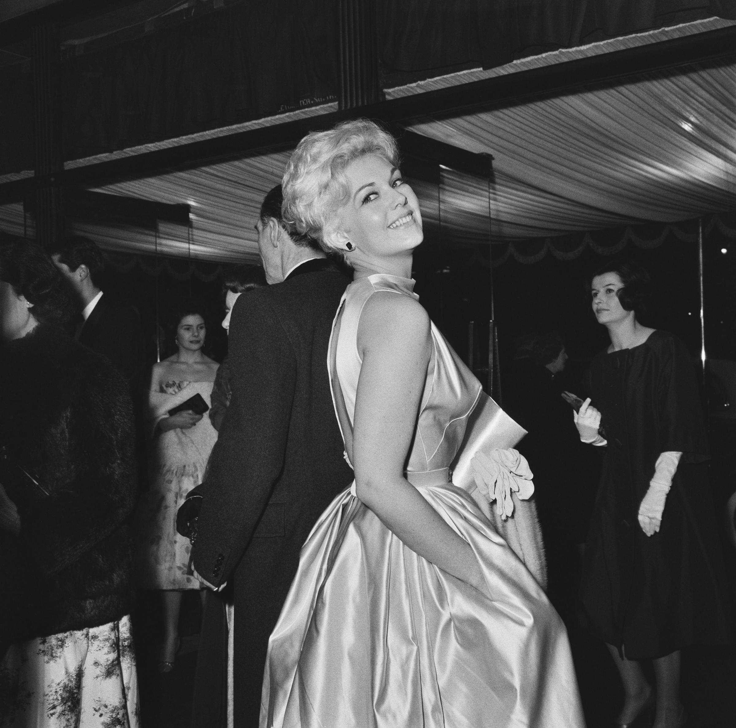 Kim Novak attends the royal performance of Once More, with Feeling at London's Odeon Leicester Square, wearing a sleeveless satin gown and smiling over her shoulder. | Source: Getty Images