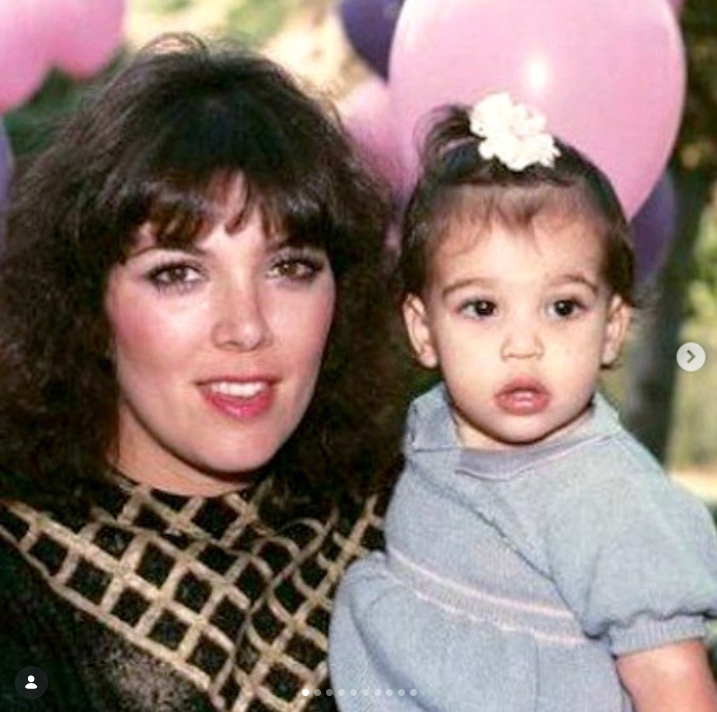 A young Kris Jenner poses with her daughter Kourtney Kardashian as a baby, holding her close while standing in front of a backdrop of pink balloons. Kris wears a patterned top, while Kourtney wears a light gray outfit with a small white flower in her hair. | Source: Instagram/krisjenner