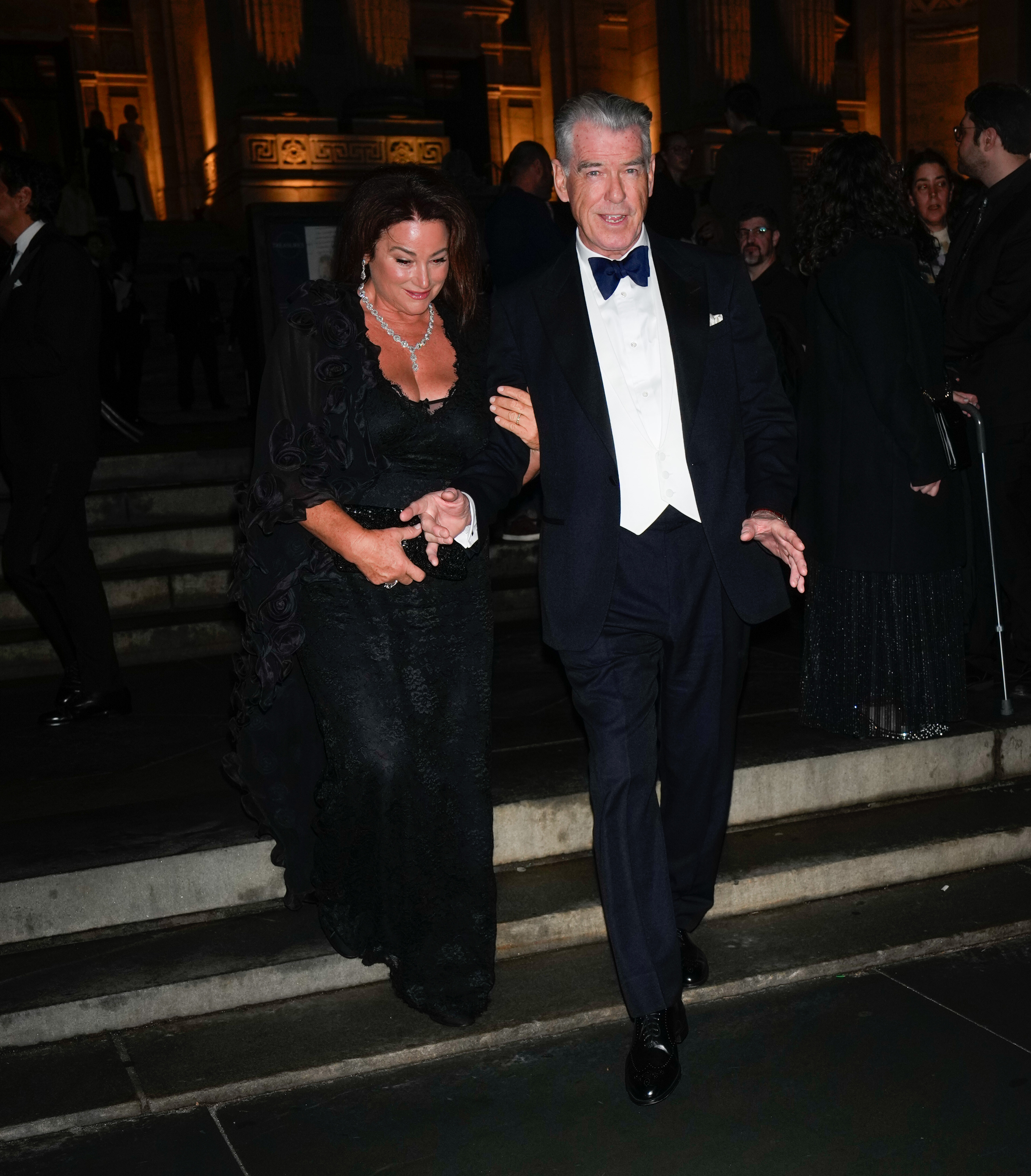 Pierce Brosnan gently holds Keely Shaye Smith's hand as they walk down the steps together, with Keely wearing an elegant black lace gown and sparkling jewelry. | Source: Getty Images.