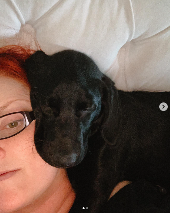 A black puppy rests its head against Wynonna's face as she lies on a white cushion. The close-up offers another glimpse of her time with animals, away from the larger outdoor spaces shown in the farm videos. | Source: Instagram/wynonnajudd