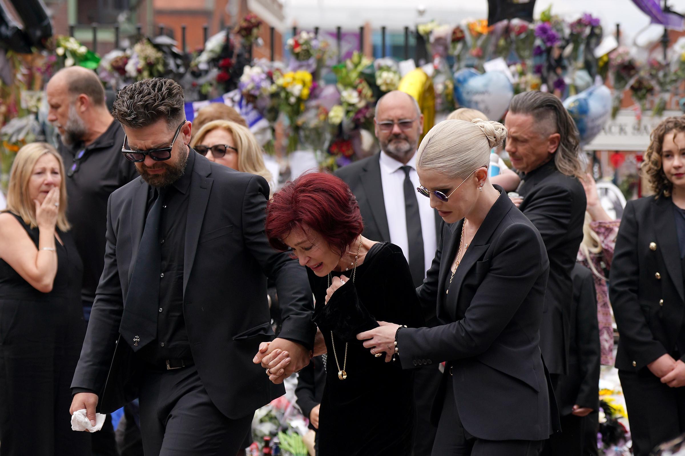 Jack, Sharon, and Kelly Osbourne move slowly together as they hold hands while approaching the messages and floral tributes left at the Black Sabbath Bench and Bridge on Broad Street in Birmingham on July 30, 2025. | Source: Getty Images