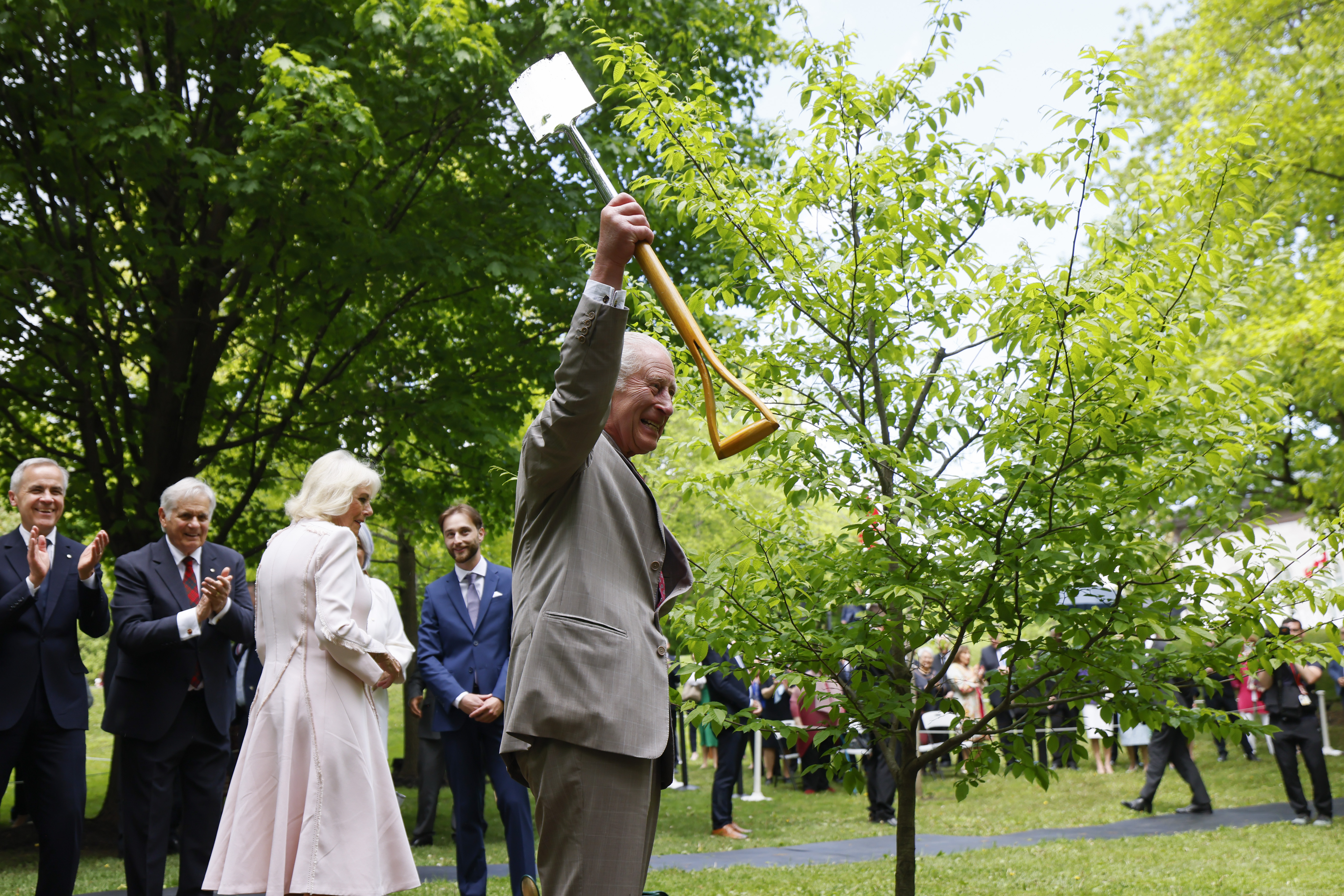 King Charles III holds a shovel as he joins Queen Camilla in a ceremonial tree planting at Rideau Hall. Surrounding them, officials—including Prime Minister Mark Carney—applaud warmly after the tree is planted, marking a moment of celebration and symbolism.