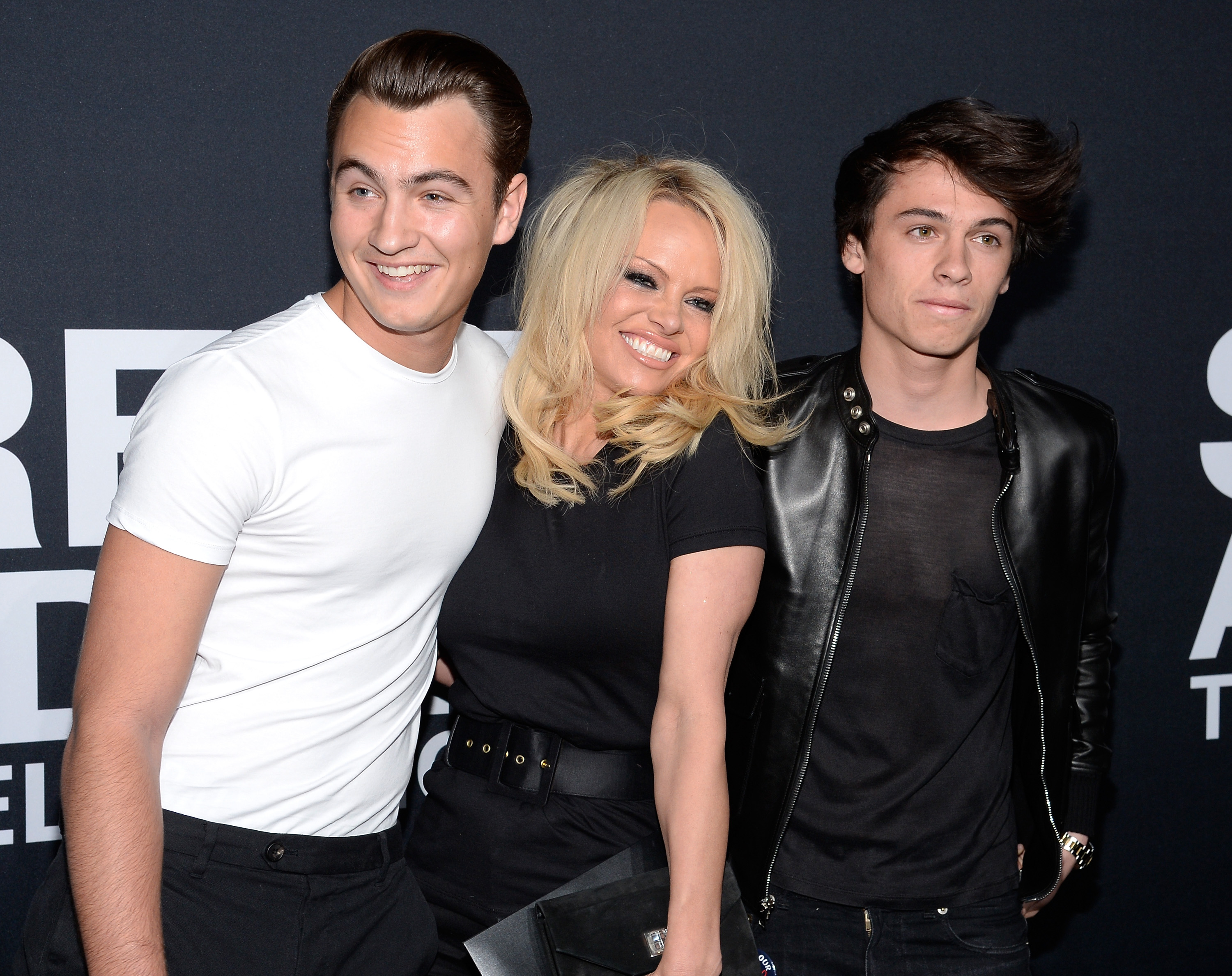 Pamela beams as she poses with Brandon and Dylan, at a 2016 red carpet event. Wearing a fitted black dress cinched with a wide belt, Anderson stands between her sons — Brandon in a crisp white T-shirt and black trousers, and Dylan in a black leather jacket layered over a sheer shirt.