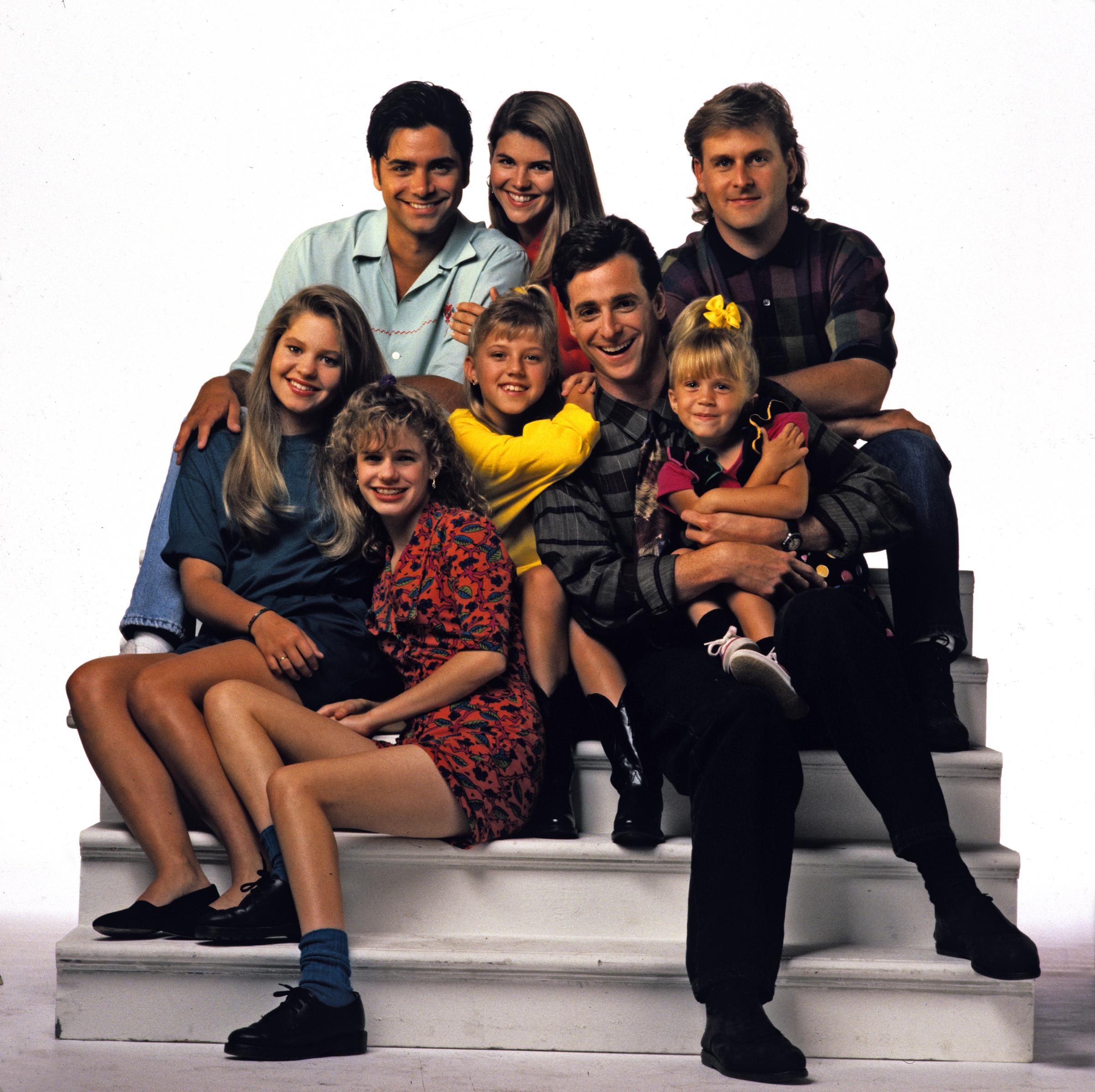 Candace Cameron Bure, Jodie Sweetin, John Stamos, and Lori Loughlin gather on white studio steps alongside Bob Saget, Dave Coulier, Andrea Barber, and Mary-Kate Olsen for a Season 5 promotional portrait on September 3, 1991. Sweetin and Cameron Bure showcase popular early '90s teen styles while Stamos and Loughlin smile from the back row, capturing the cast at the peak of "Full House" as their lovable family dynamic continued to dominate Friday night television.