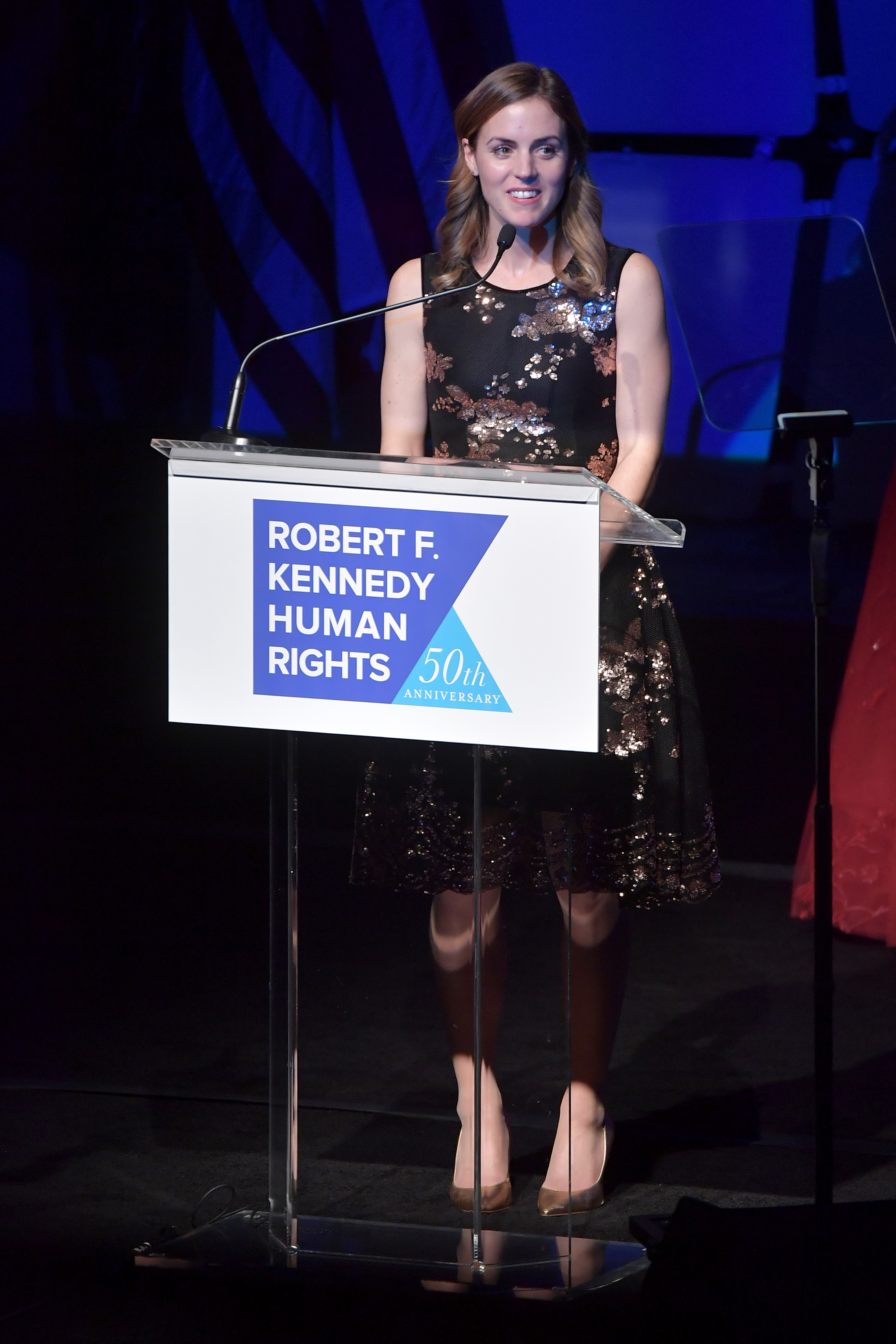 A poised Kyle Kennedy Kerr pauses with a subtle smile while presenting at the 2019 Robert F. Kennedy Human Rights Ripple of Hope Awards on December 12, 2018, in New York City. Captured from a slightly elevated vantage behind the branded 50th-anniversary lectern, she lights up the stage in a floral-patterned sequin cocktail dress with a flared knee-length skirt and pointed metallic heels against a deep blue backdrop. | Source: Getty Images