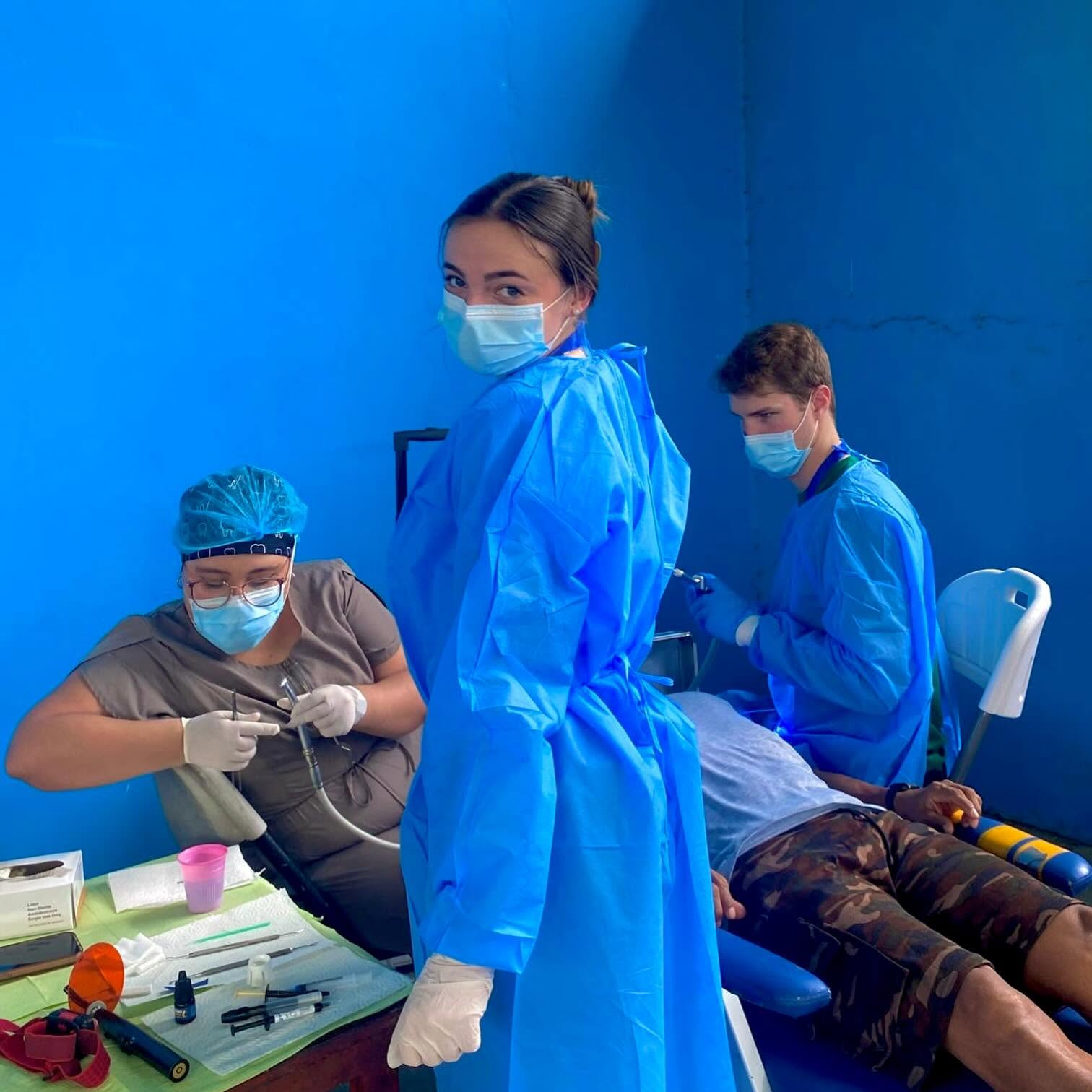 Leah Victoria Fonte glances over her shoulder while volunteering on a dental mission brigade in Tortí, Panama. Dressed in full protective gear, including a bright blue surgical gown, a medical face mask, and disposable gloves, she stands in a field clinic as fellow volunteers tend to a patient seated in a mobile dental chair. | Source: Facebook/Leah Fonte
