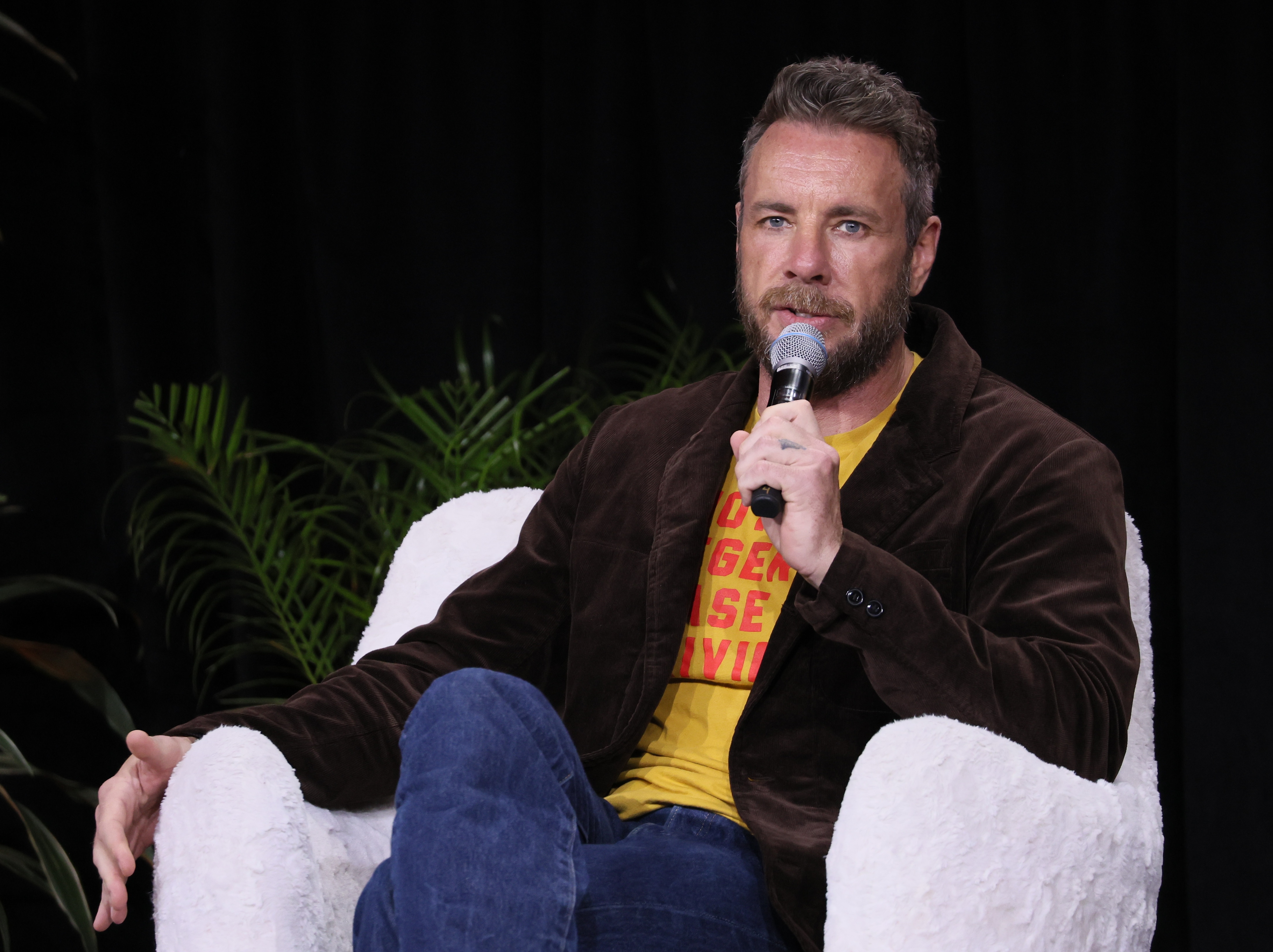 Dax Shepard speaks during a panel at the Natural Products Expo West, seated comfortably in a white chair. Dressed in a yellow graphic tee and brown jacket, he shares insights with relaxed energy and quiet authority.