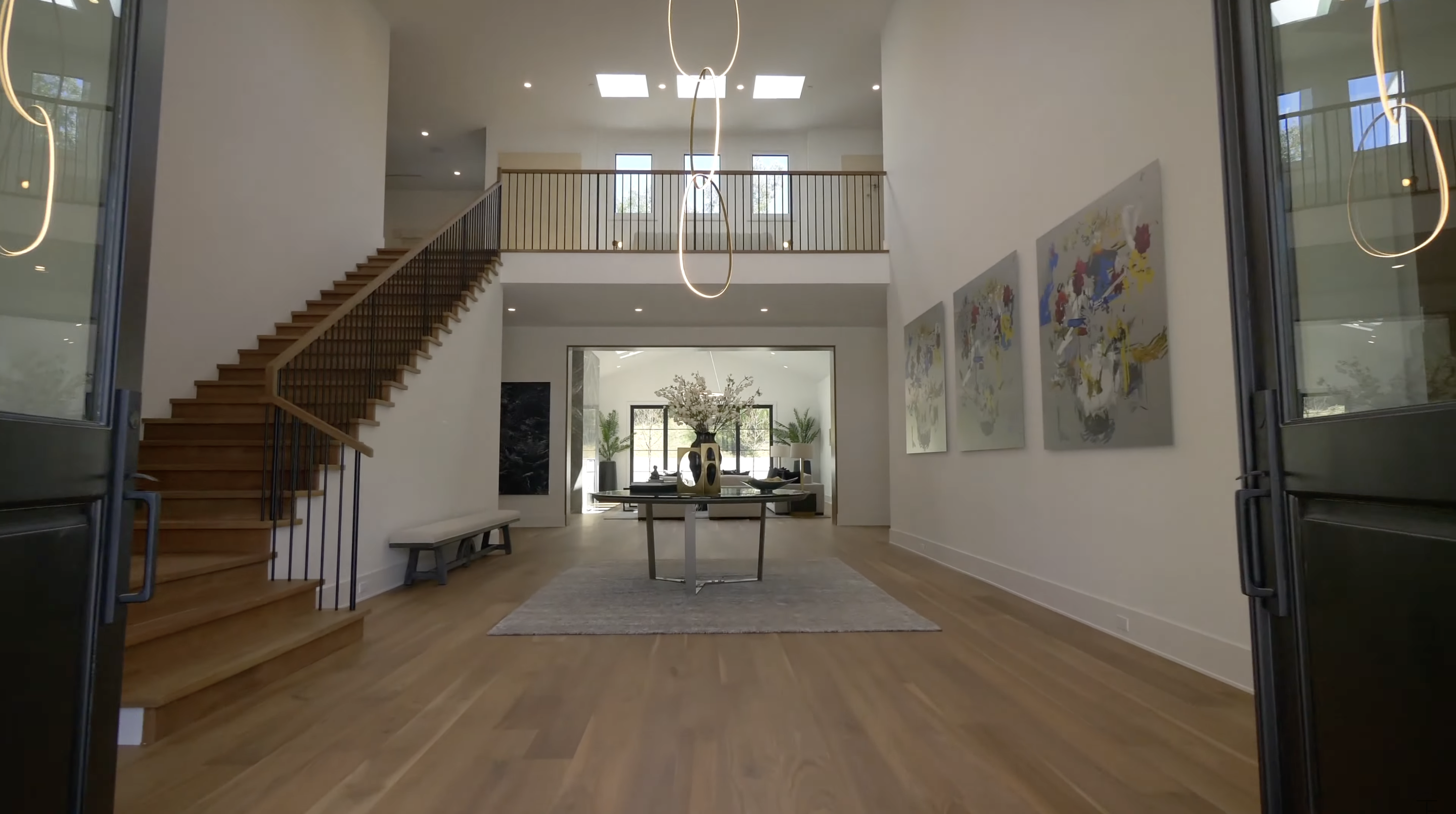 Warm wood floors run through the tall entry, while slender black railings define the staircase and upper level. Looping light fixtures draw attention toward the skylights overhead. | Source: Youtube/Tomer.Fridman