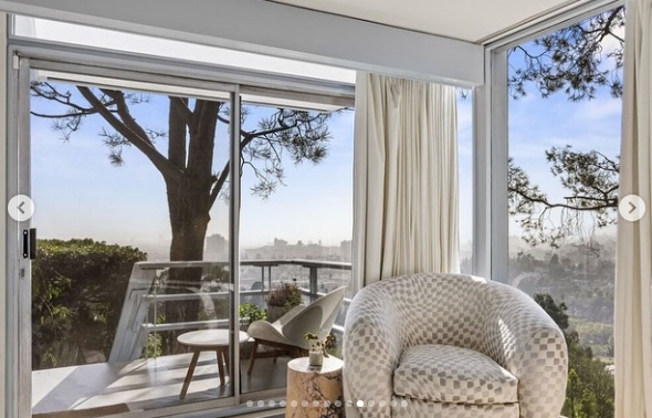A quiet sitting corner takes full advantage of the home's hillside position, pairing a patterned armchair with glass walls and doors opening onto a small terrace. Mature trees rise immediately outside, while distant city views stretch beyond them, turning the intimate nook into another light-filled vantage point. | Source: Instagram/jparkerstanton