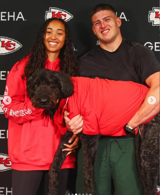 George Karlaftis and Kaia Harris pose together in front of a Kansas City Chiefs backdrop while holding a large black dog dressed in a red hoodie. | Source: Instagram/chiefs and nfl