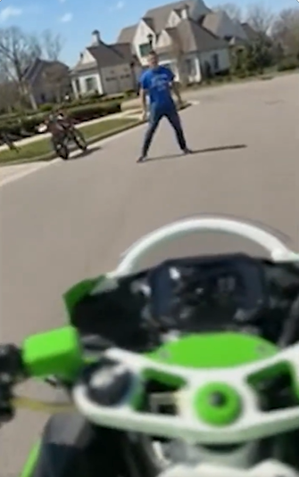 Ronnie Taylor in a blue shirt stands in the middle of the street with legs apart, blocking the path as Alan Ritchson's motorcycle approaches. | Source: X/TMZ