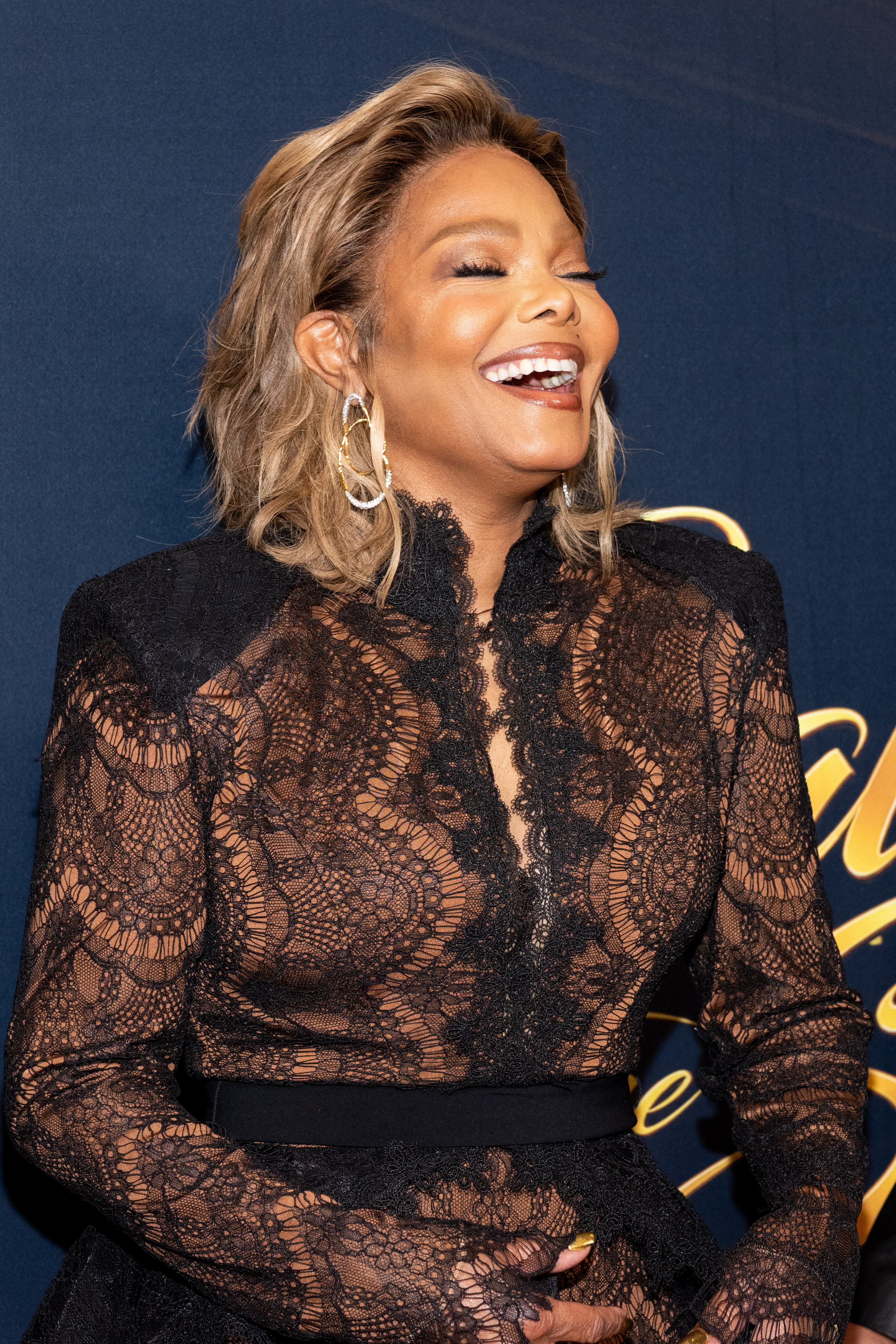 Janet Jackson laughs with her eyes closed as she poses in her sheer black lace gown, showcasing its scalloped neckline, structured shoulders, and wide black waist belt. | Source: Getty Images