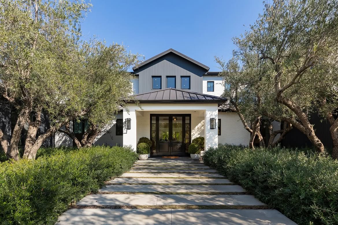 The later exterior presents a more restrained combination of white masonry, dark trim and gray vertical siding. Mature olive trees and dense plantings frame the walkway, partially screening the entrance from view. | Source: Instagram/theshevins