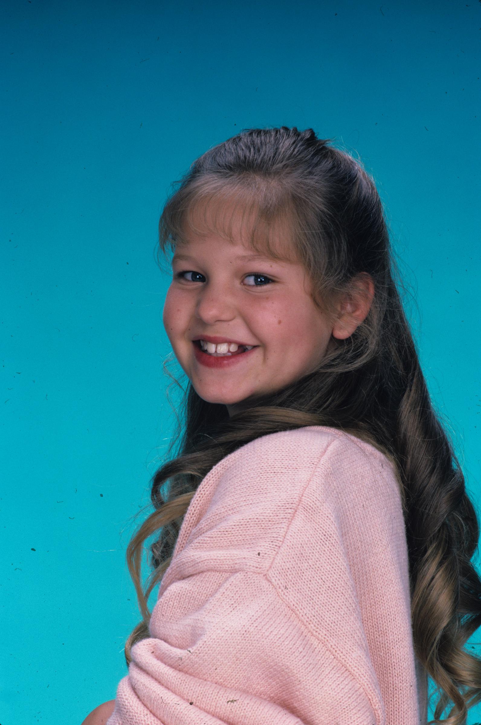 Young Candace Cameron smiles brightly during a promotional portrait session for the pilot episode "Our Very First Show" of the hit ABC sitcom "Full House" on September 22, 1987, where she starred as eldest daughter D.J. Tanner. Posing against a vibrant turquoise backdrop, the child actress wears a pastel pink knit sweater with her long brown hair styled in soft curls and finished with classic straight fringe. | Source: Getty Images