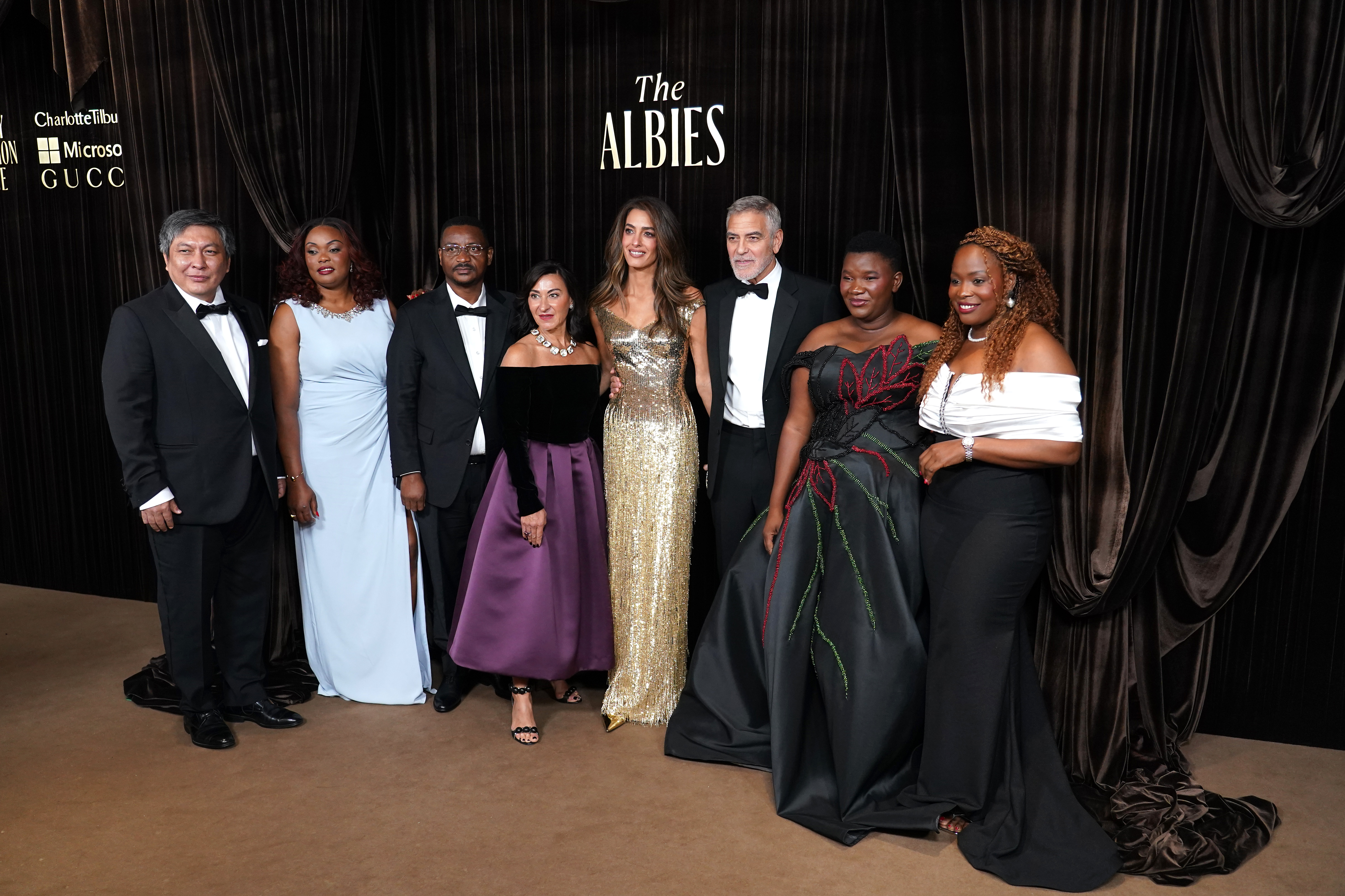 George Clooney and Amal Clooney pose with fellow guests and honorees at the Clooney Foundation for Justice's 2026 Albies at London's Natural History Museum on October 9. The group stands before a black-curtained backdrop bearing The Albies logo, dressed in formal eveningwear, with several guests displaying their floor-length gowns. | Source: Getty Images