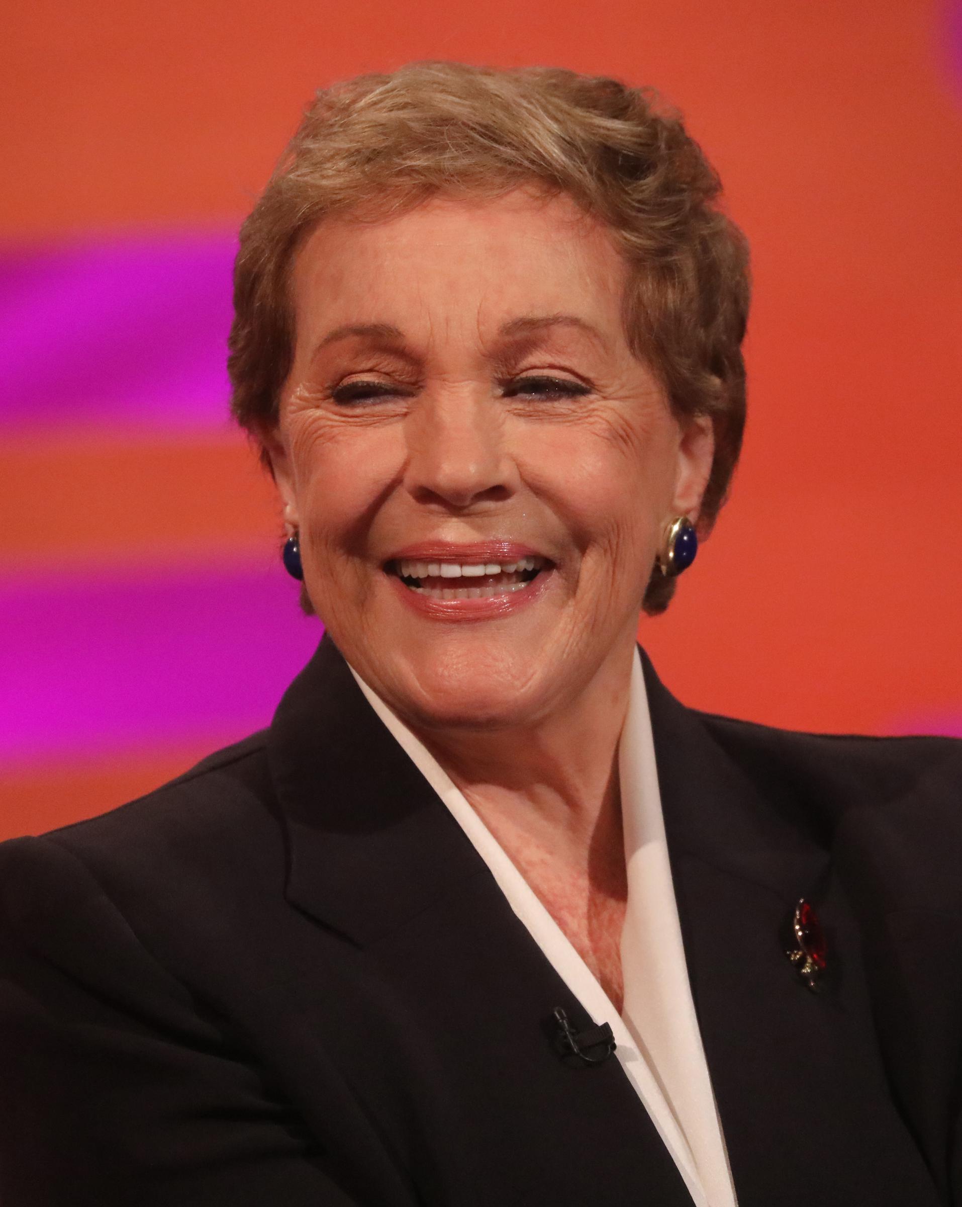 Julie Andrews breaks into a joyful laugh during the filming of "The Graham Norton Show" at BBC Studioworks 6 Television Centre in London on October 31, 2019. The legendary actress looks effortlessly sharp in a tailored black suit jacket over a crisp white blouse, accented with a remembrance poppy pin on her lapel and bold royal-blue and gold cabochon clip earrings.