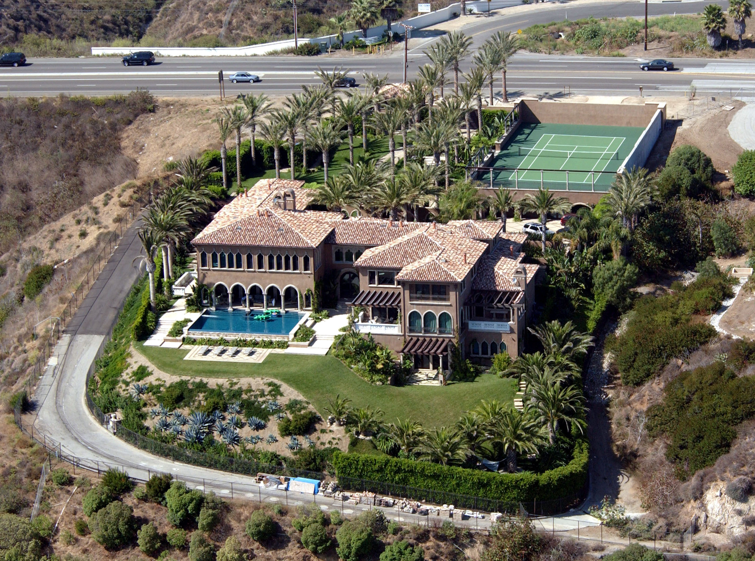 An aerial view captured Cher's sprawling Italian Renaissance-style estate in Malibu, California, on August 28, 2003. The property showcases terracotta-tiled roofs, an infinity-edge swimming pool overlooking manicured lawns, a private tennis court flanked by tall palm trees, and the winding hillside access road positioned just below the Pacific Coast Highway. | Source: Getty Images