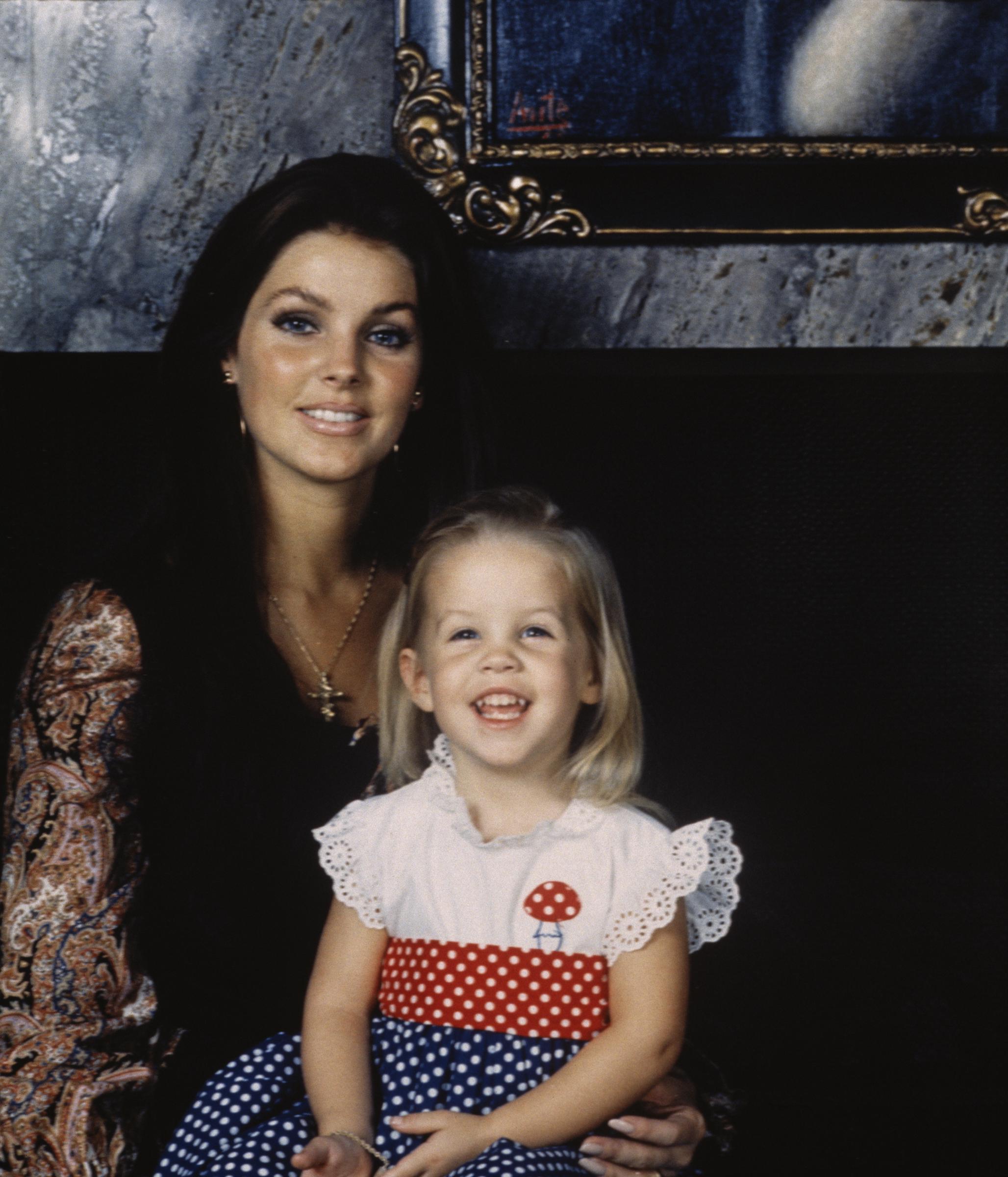 Priscilla Presley poses with her toddler daughter, Lisa-Marie Presley, during Thanksgiving week in November 1970 at their home on Hillcrest Road in Beverly Hills, California, wearing a dark top with colorful paisley sleeves and a gold cross necklace, while Lisa-Marie smiles in a white eyelet-trimmed polka-dot dress with a red mushroom motif. | Source: Getty Images