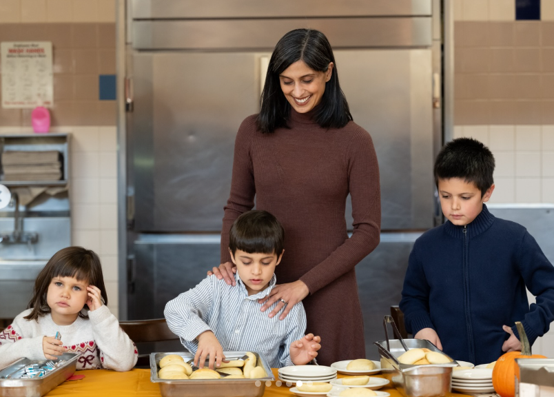 Usha stands behind her children at the serving table, gently guiding them as they select bread rolls and plates, reinforcing the communal and hands-on nature of the event. | Source: Instagram/vp
