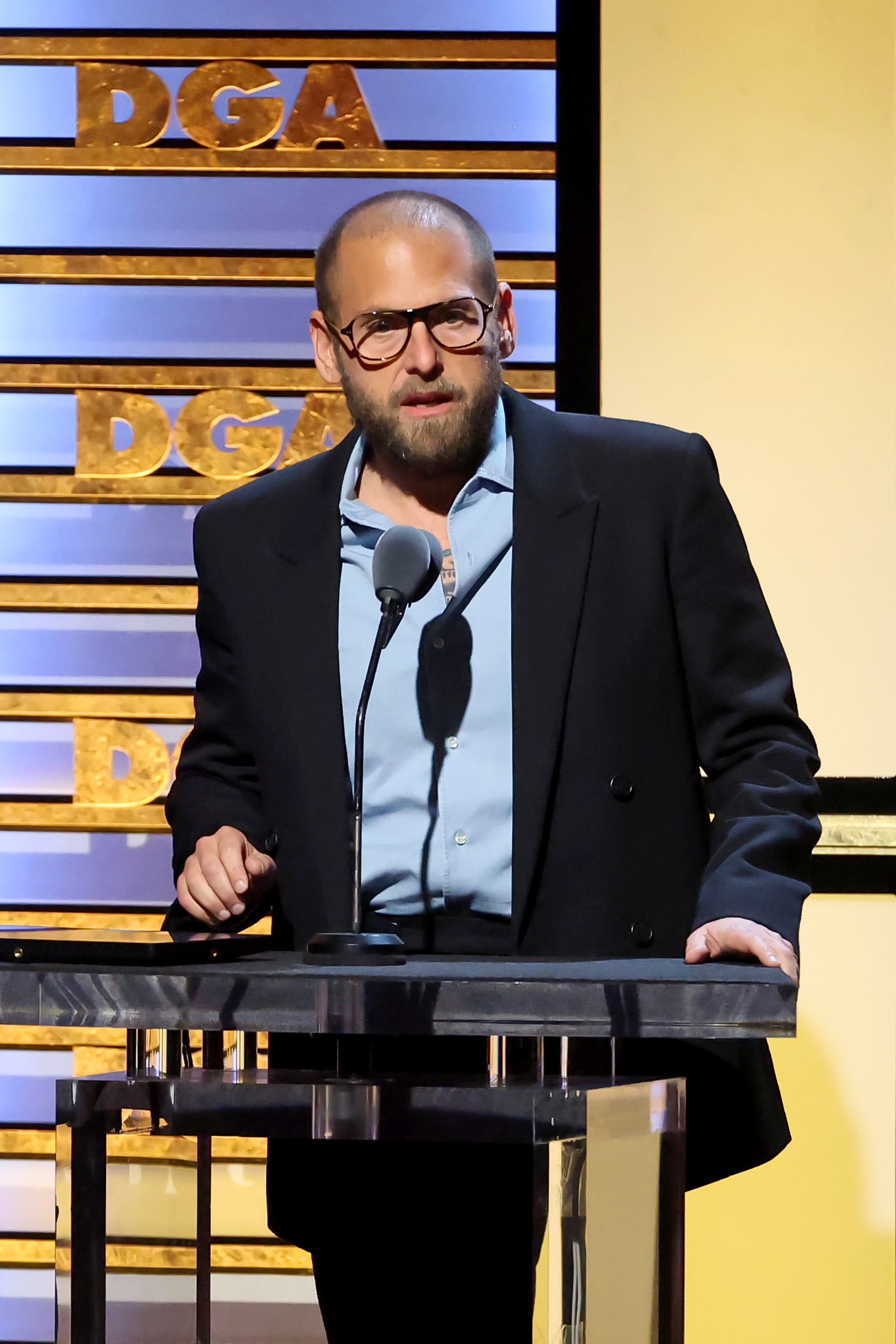 Jonah Hill took the stage during the 76th Directors Guild of America Awards at The Beverly Hilton in Beverly Hills, California, on February 10, 2024. Wearing a dark oversized jacket over a light blue shirt, Hill appeared with a closely shaved head, neatly trimmed beard, and statement glasses as he addressed the audience from the podium. His notably leaner frame marked another chapter in the actor's long-running physical transformation.