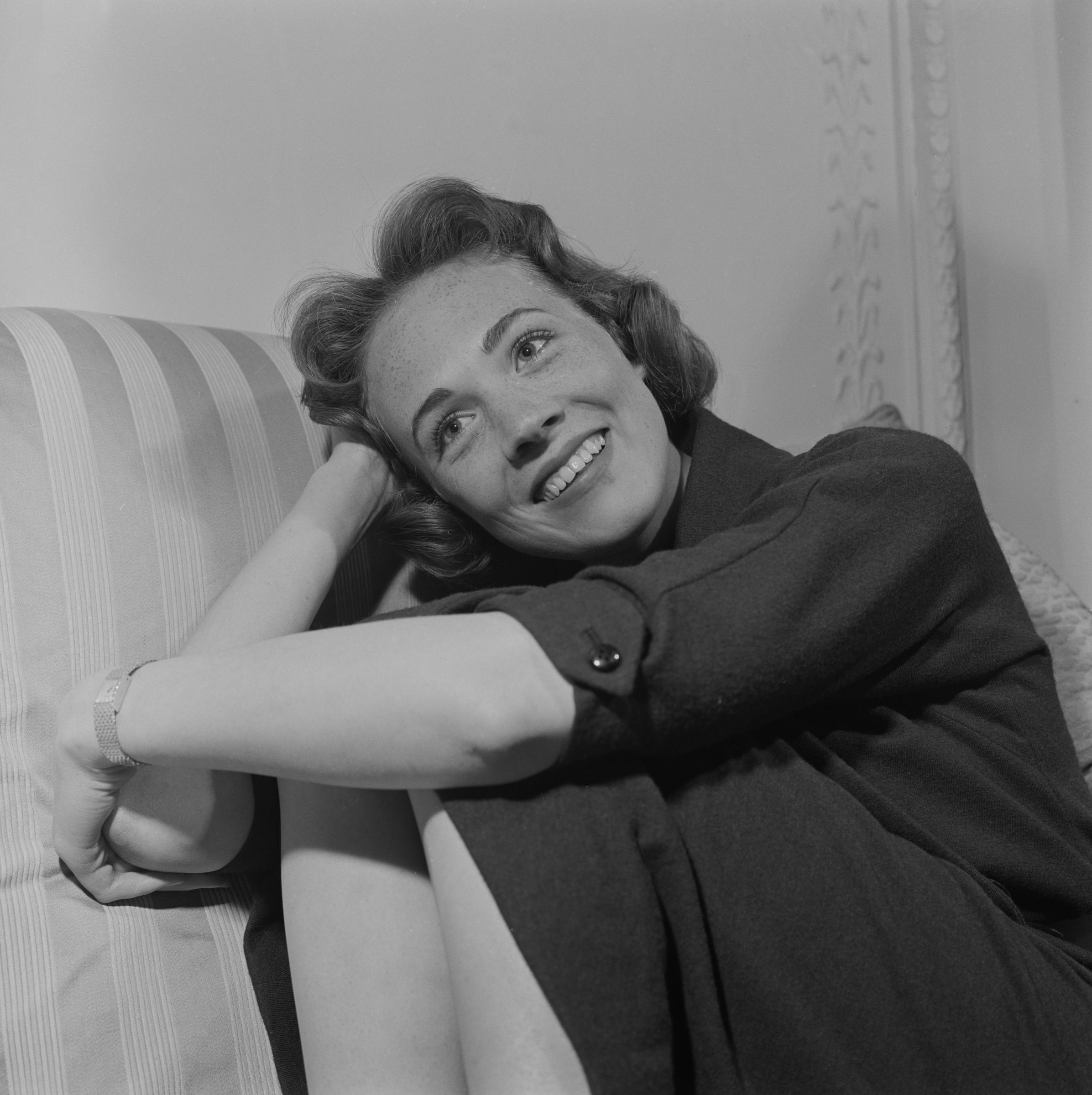 A candid portrait captures Julie Andrews relaxing on an armchair on April 17, 1958, just weeks before reprising her acclaimed role as Eliza Doolittle in the West End production of "My Fair Lady" at London's Theatre Royal, Drury Lane. The actress smiles warmly with her head propped against her hand, wearing a tailored dark three-quarter-sleeve dress and a classic wristwatch, her styled brunette curls framing her joyful expression.