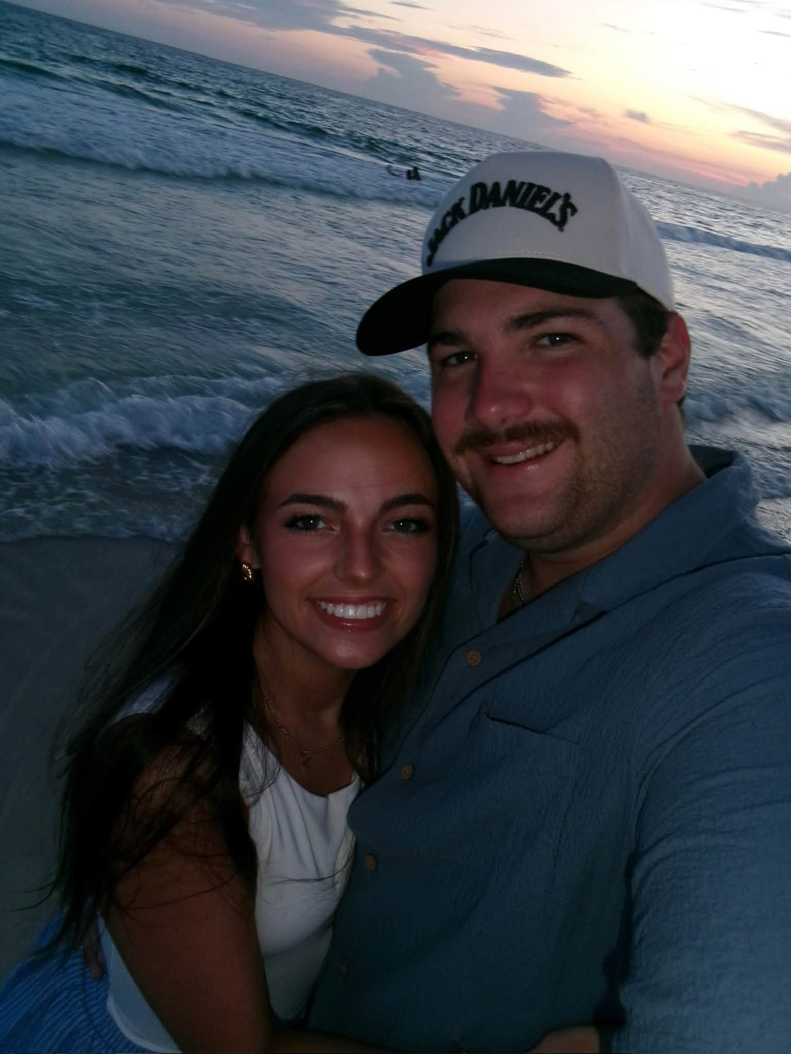 Leah Victoria Fonte and Shane David Hirstius smile closely together for a beach selfie at sunset. Leah leans gently against Shane, wearing a white top and dainty gold jewelry, while Shane smiles warmly in a blue button-down shirt and a two-tone baseball cap with ocean waves and a dusk sky softly visible behind them. | Source: Facebook/StevenMarjorie Hirstius