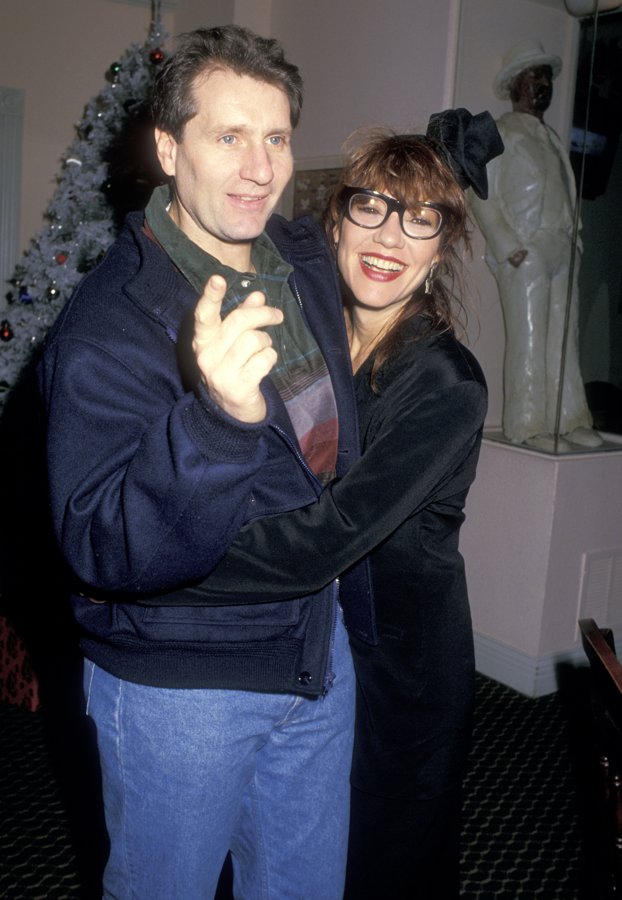 Ed O'Neill and Katey Sagal shared a cheerful, affectionate embrace while attending the Feed the Valley Homeless Benefit on December 13, 1987, at the Sherman Oaks Galleria in Sherman Oaks, California. Posing beside a decorated holiday tree, the "Married... with Children" co-stars brought playful chemistry to the festive charity event, with O'Neill pointing mid-laugh in a navy bomber jacket and denim jeans as Sagal wrapped both arms around him, flashing a wide smile in thick-rimmed glasses, a black blazer dress, and a whimsical mini top-hat fascinator. | Source: Getty Images