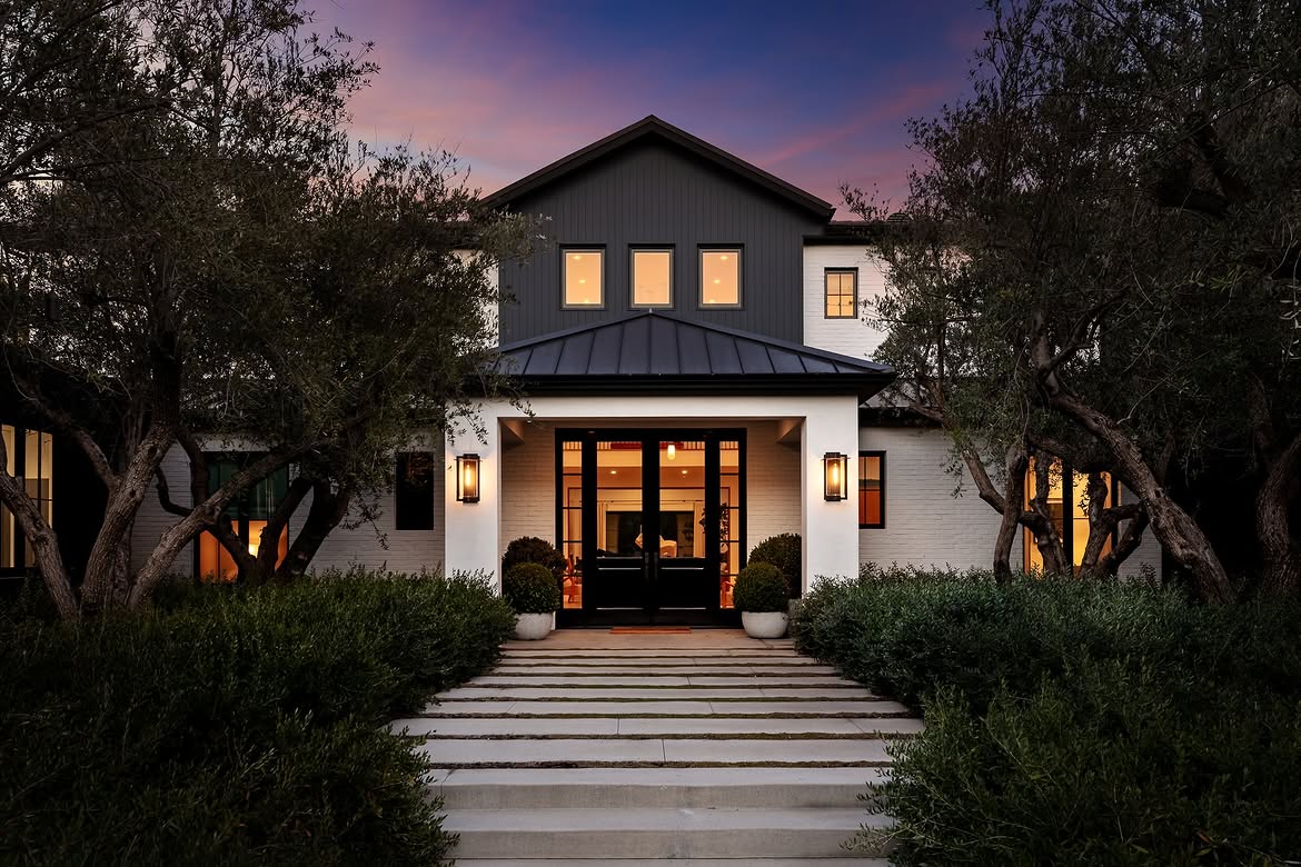 At dusk, warm interior lighting changes the appearance of the home's restrained white, gray, and black façade. Olive trees and thick landscaping continue to frame the approach, while the illuminated entrance becomes the focal point. | Source: Instagram/tomer.fridman