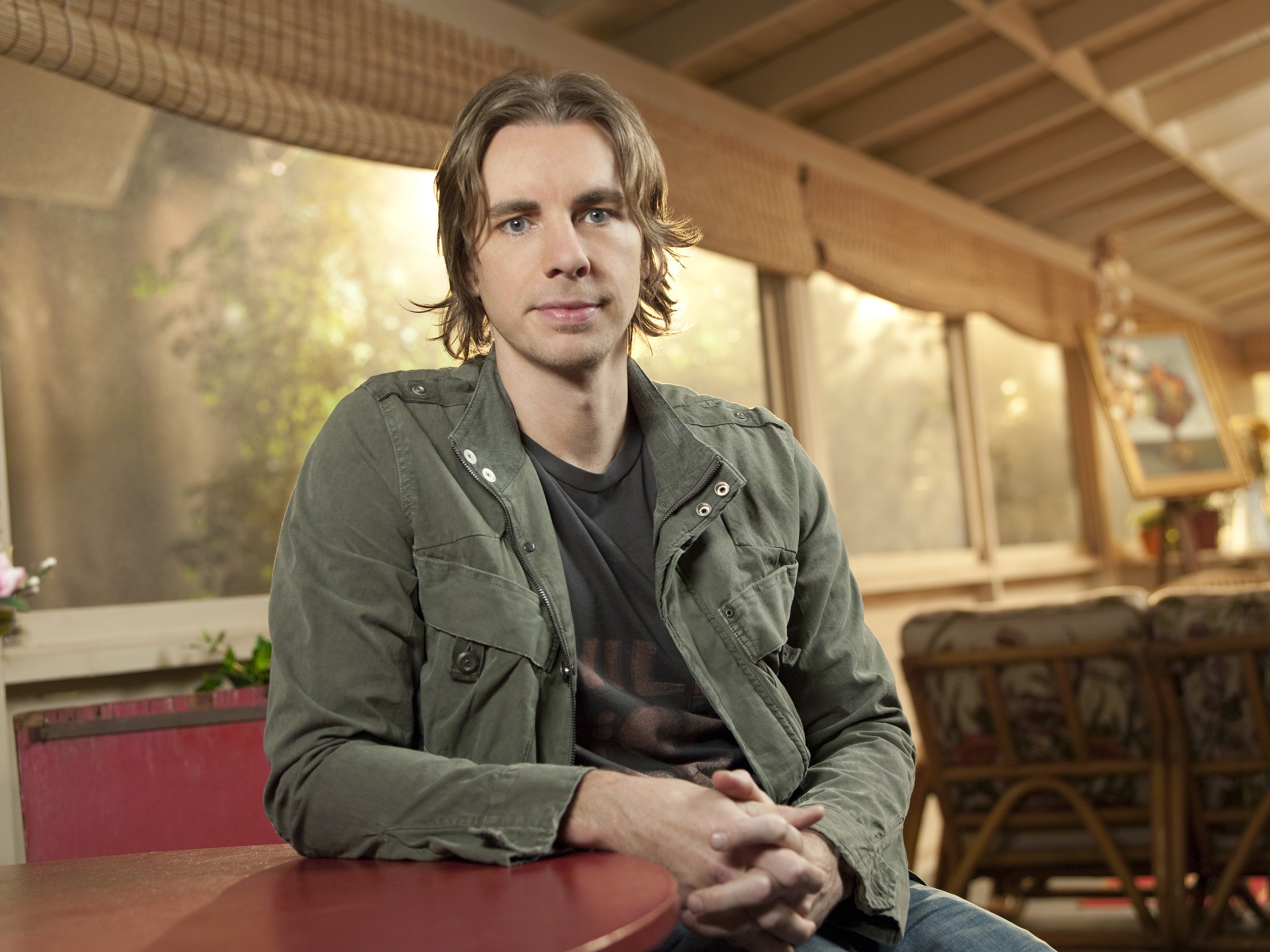 Dax Shepard sits at a rustic table, dressed in a green jacket with longer, tousled hair. His laid-back style and thoughtful gaze reflect his character's calm confidence in a dramatic role.