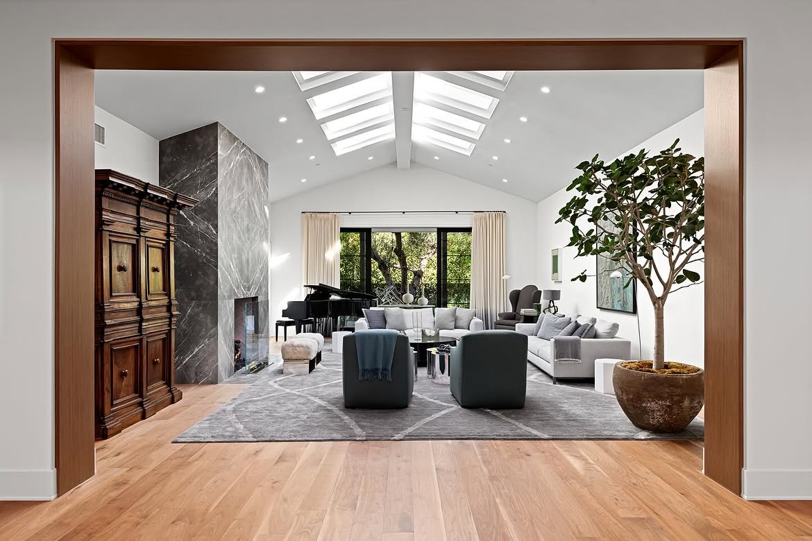 A dark stone fireplace rises toward the vaulted ceiling, providing a dramatic contrast to the white walls and pale seating. A grand piano, large indoor tree and mix of contemporary and traditional pieces give the room more visual variety. | Source: Instagram/theshevins
