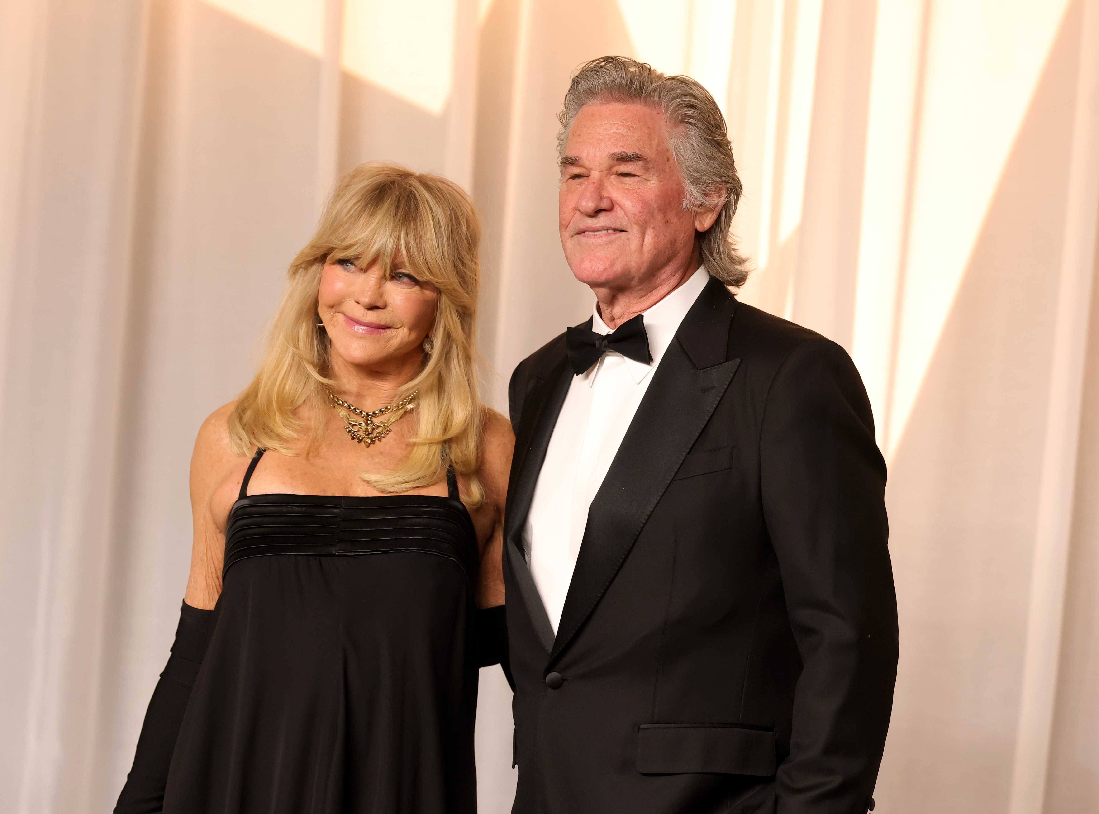 Hawn and Russell pose together in elegant black-tie looks, with Hawn's strappy gown, long gloves, statement jewelry, and voluminous blonde hair contrasting with Russell's tailored tuxedo, crisp white shirt, and swept-back gray hair.