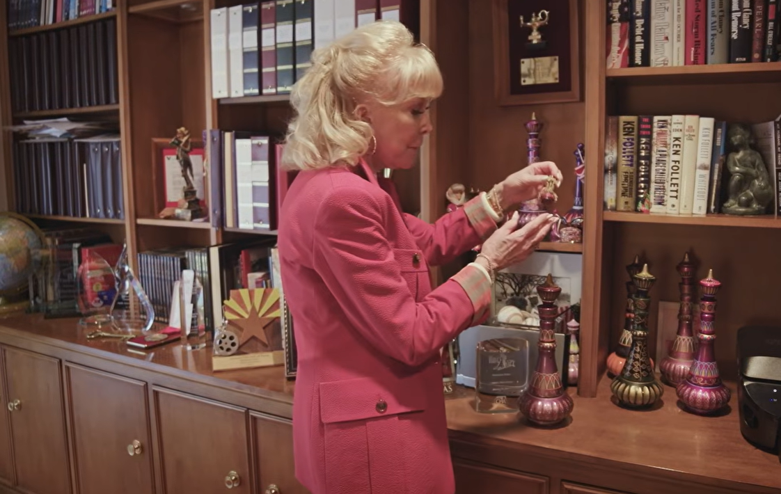 Barbara Eden examines a small decorative collectible in her home office, surrounded by colorful genie bottles, books, and career memorabilia displayed across her wooden shelves. | Source: YouTube/People