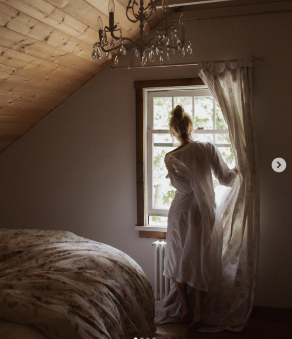 Viewed from behind in an ethereal white nightgown with her hair swept in a topknot, Pamela Anderson pulls back a patterned drape to look out a sunlit dormer window at Arcady. Bathed in soft morning light beneath a crystal chandelier and sloped wood-plank ceiling, the cozy attic bedroom perfectly mirrors her caption: "Fall is a beautiful time of year… restful, peaceful… protective, fuzzy socks… cocooning, hibernating… 🤍". | Source: Instagram/pamelaanderson