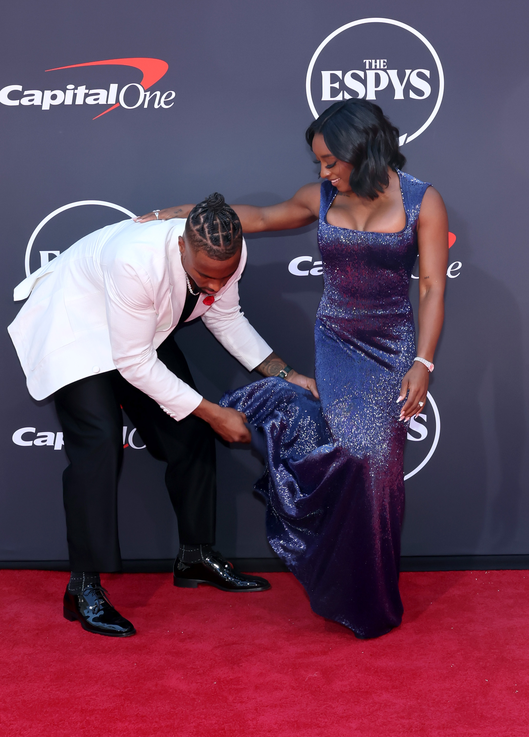 Jonathan Owens helps adjust Simone Biles' dress as they pose together on the red carpet. Biles smiles down at him while holding his shoulder for balance, as he lifts the hem of her sparkling purple gown to prevent it from dragging. The moment captures a candid display of teamwork and care between the couple.