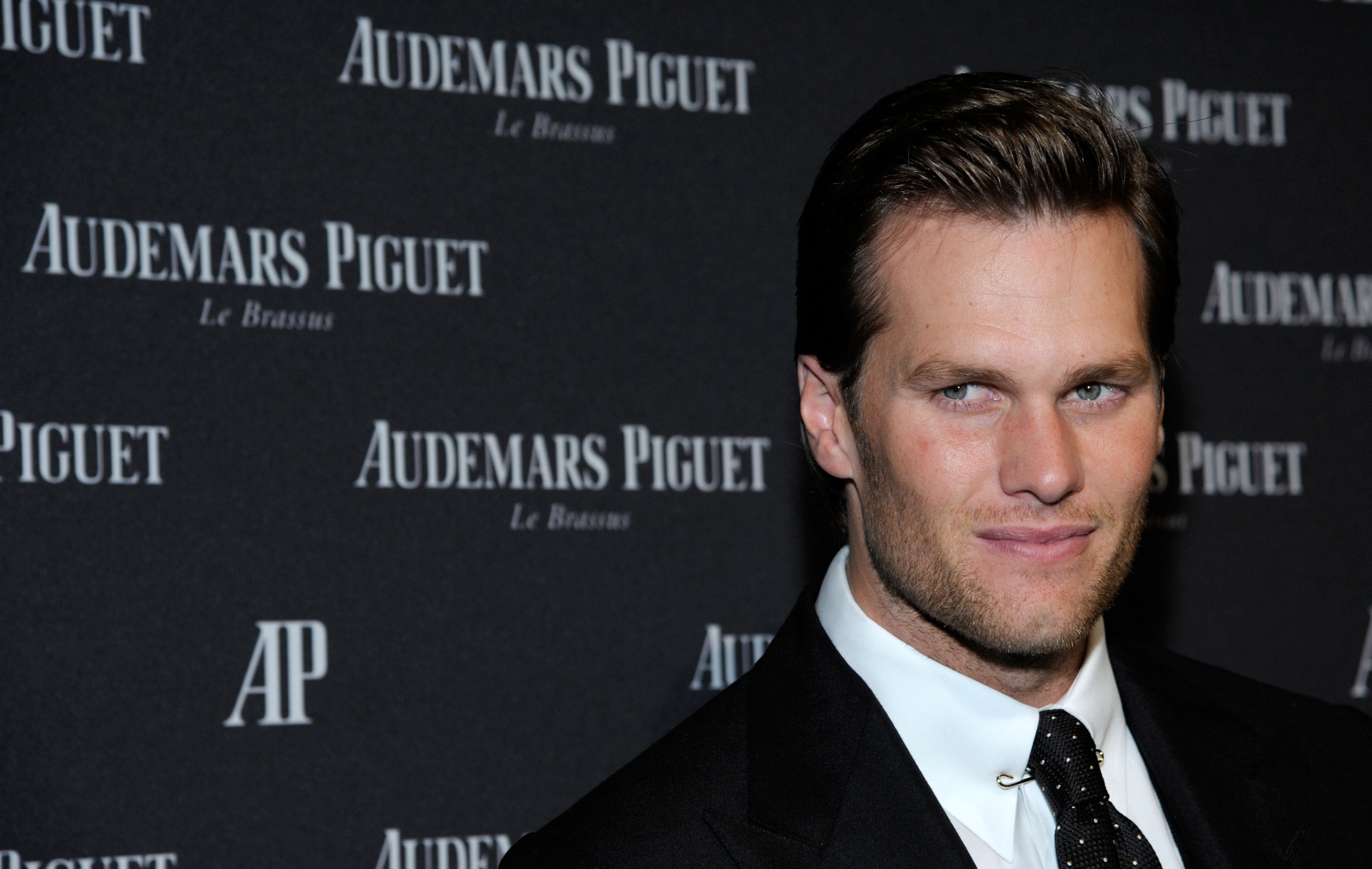Tom Brady attends the Royal Oak 40 Years: From Avant-Garde to Icon event at the Park Avenue Armory in New York City. He is dressed in a sleek black suit paired with a crisp white shirt and a black-and-white polka-dot tie. His jawline looks perfectly defined as he smiles slightly.