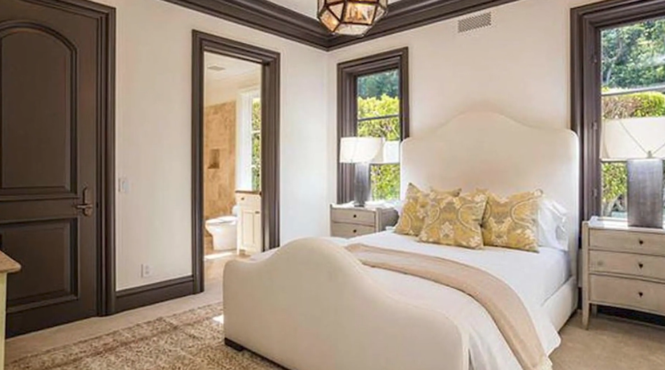 An airy secondary bedroom combines crisp neutral bedding and an upholstered camelback headboard with bold espresso-stained moldings, framed garden views, and a dedicated en suite bathroom. | Source: YouTube/Taste of Country
