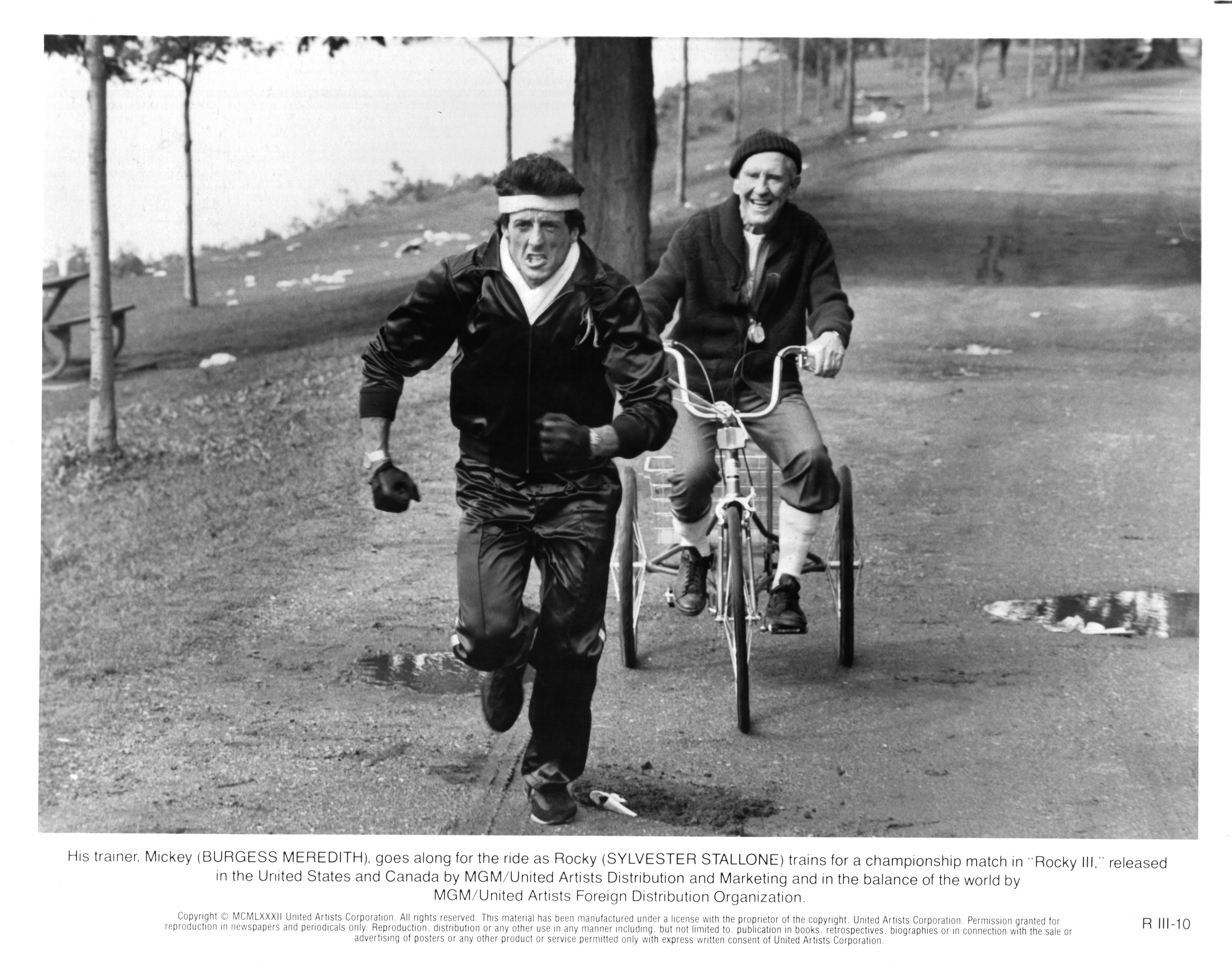 Sylvester Stallone charges uphill as Rocky Balboa while Burgess Meredith follows closely behind on a bicycle as trainer Mickey Goldmill in a scene from "Rocky III." The training sequence reflects the demanding conditioning behind Rocky's preparation, with Stallone seeking a leaner, faster version of the boxer for his showdown with Clubber Lang.