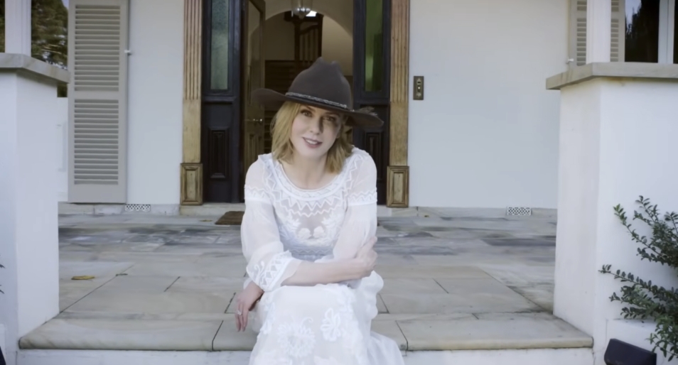 Dressed in white with a wide-brimmed hat, Nicole Kidman sits at the front steps of the farmhouse, highlighting the home’s elegant facade with shuttered windows and carved wood accents. From a video dated July 20, 2015. | Source: YouTube/Vogue