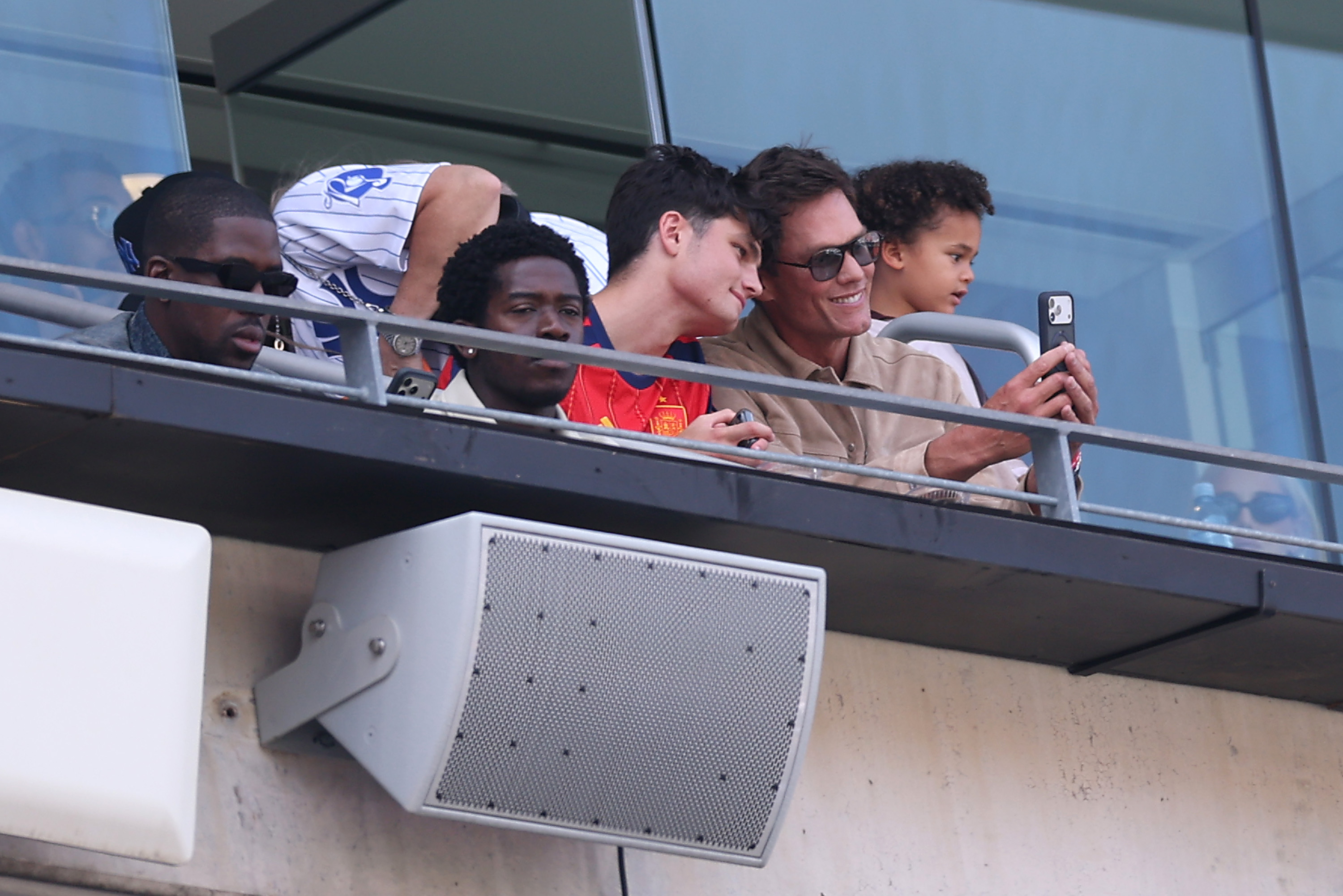 Tom Brady smiles and captures a selfie with his young son peering over his shoulder from their stadium balcony suite, enjoying football's biggest night at the FIFA World Cup 2026 Final match. | Source: Getty Images