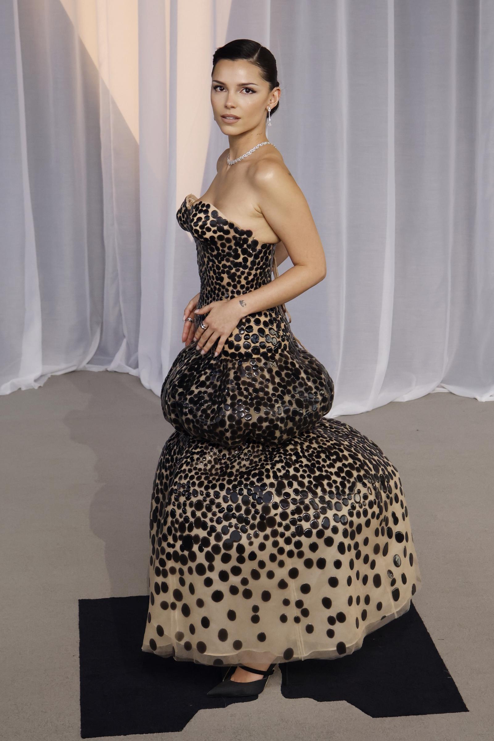 Inde Navarrette brings striking, avant-garde drama in a sculpted, strapless gown patterned with glossy, oversized polka dots over a neutral underlay. The voluminous silhouette flares boldly at the hips and tiered bubble hem, which she balances with a sleek, polished updo, a sparkling diamond choker, and sharp black pointed-toe heels.