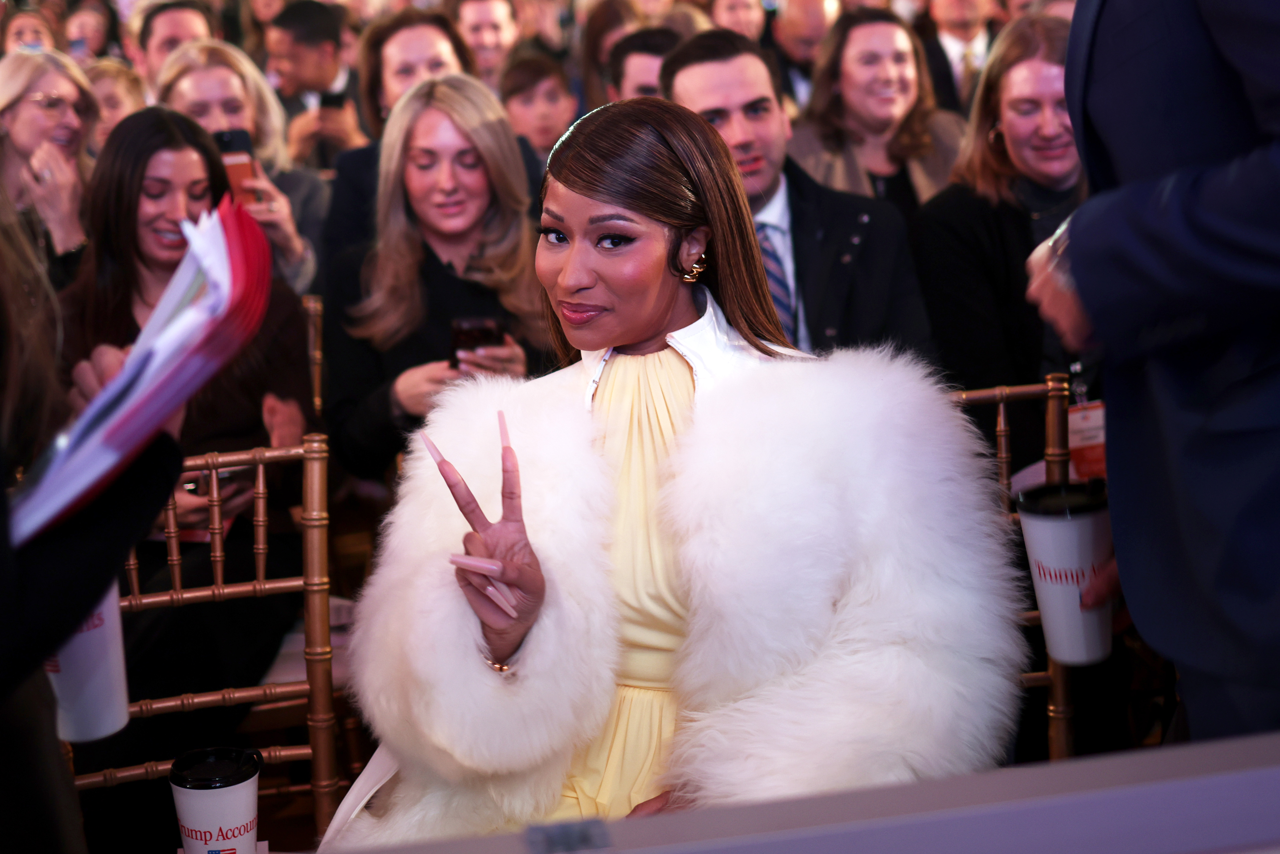 Nicki Minaj flashes a peace sign while seated at a Trump Accounts event, wearing a voluminous white fur coat over a pale-yellow outfit. The appearance offered another example of her mixing glamorous fashion with more formal public occasions. | Source: Getty Images
