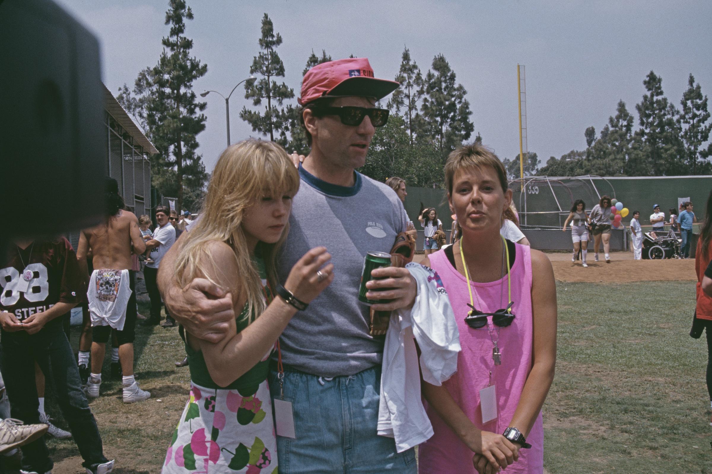 Christina Applegate, Ed O'Neill, and Amanda Bearse hung out outdoors at a charity baseball game circa 1990 in Los Angeles, California. The "Married... with Children" co-stars sported quintessential late-1980s and early-1990s summer styles: Applegate wore a white minidress printed with colorful umbrellas, O'Neill wrapped an arm around her while wearing a grey tee, acid-wash denim shorts, dark sunglasses, and a hot pink cap, and Bearse stood beside them in a neon pink tank top with sunglasses hanging around her neck. | Source: Getty Images