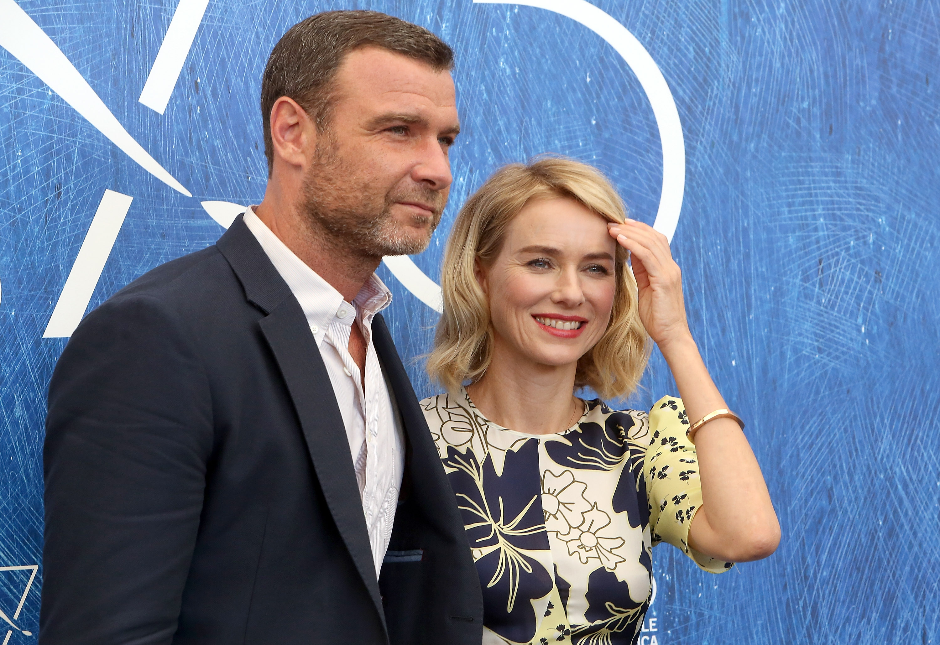 Naomi Watts and Liev Schreiber pose together at a press event against a bright blue backdrop. Watts wears a patterned dress and smiles toward the cameras. Schreiber stands beside her in a navy suit, giving the moment a polished red-carpet feel.