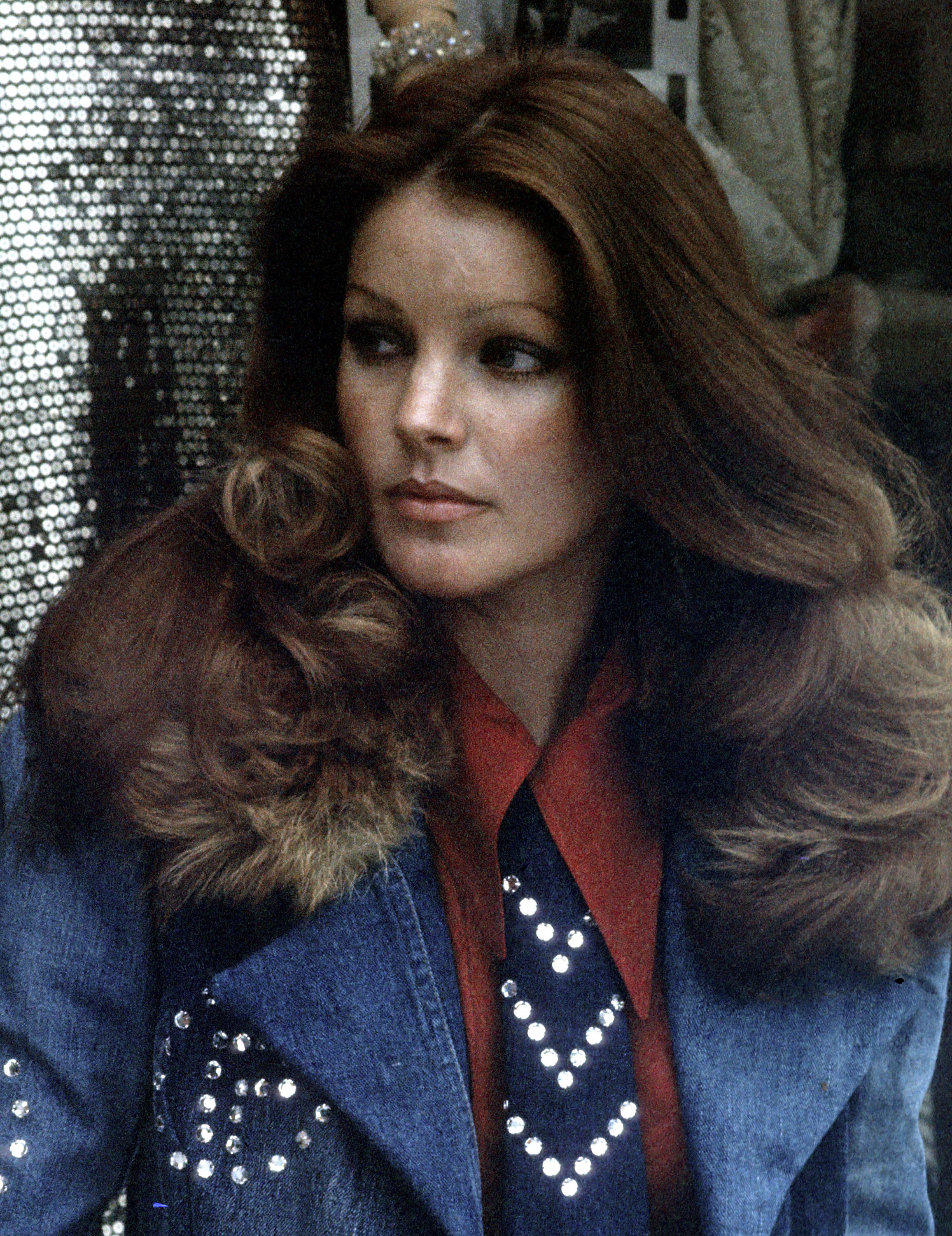 Priscilla Presley poses during a photo session at her clothing store, Bis & Beau Boutique, in Beverly Hills, California, on March 19, 1974, wearing a denim jacket with fur collar trim and rhinestone embellishments layered over a rust-red collared shirt and a studded tie, with her auburn hair styled in voluminous feathered waves.