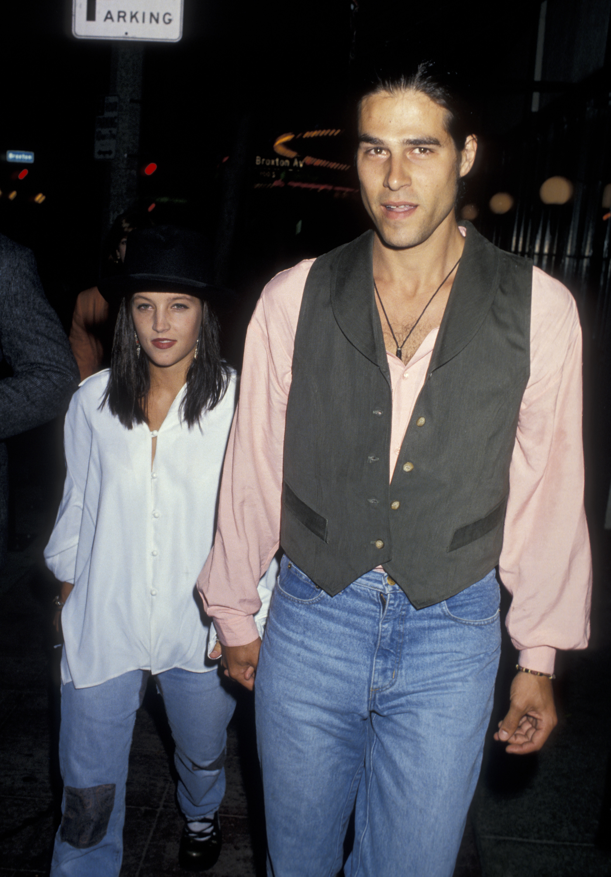 Lisa Marie Presley holds hands with her husband Danny Keough as they arrive at the Los Angeles screening of "The Naked Gun 2 1/2: The Smell of Fear" at Mann's Bruin Theater in Westwood, California, on June 13, 1991, with Presley dressed in an oversized white button-down blouse, light-wash jeans, and a black bowler hat, while Keough wears a pale pink collared shirt under a gray vest paired with denim jeans.