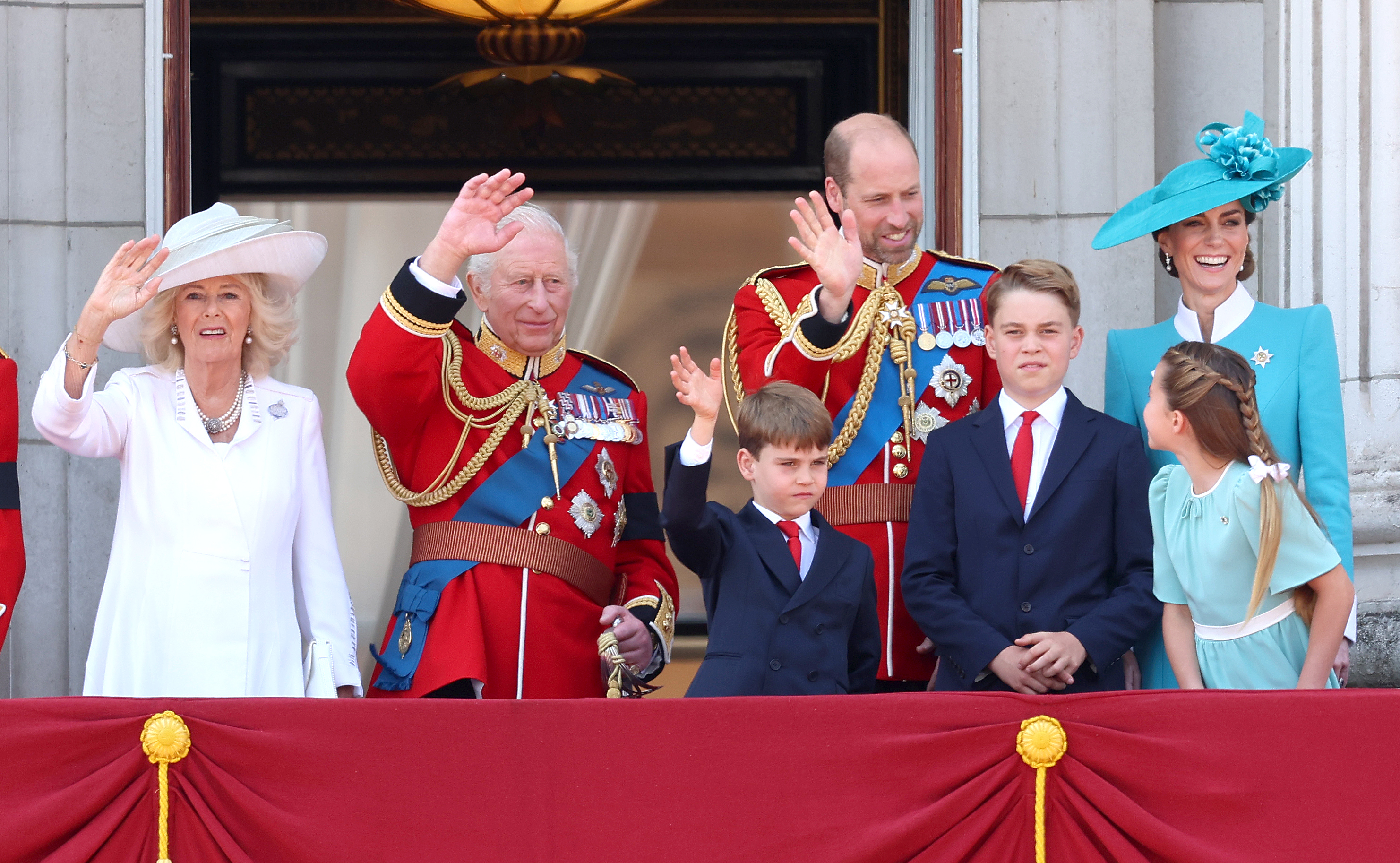 Queen Camilla, King Charles III, and the Prince and Princess of Wales appear on the Buckingham Palace balcony during Trooping the Colour 2025, joined by their children—Prince George, Princess Charlotte, and Prince Louis. In this medium shot, Princess Catherine stands out in a tailored teal blazer featuring sharp white cuffs and a structured white collar. The ensemble, visible in crisp detail, echoes regal sophistication as the royal family observes the military spectacle and waves to the gathered crowds below.