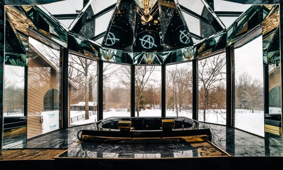 Floor-to-ceiling windows frame the snowy grounds outside a dramatic bathing area in Mike Tyson's former residence, where mirrors cover much of the angular ceiling above a sunken tub. Gold accents contrast with dark marble across the room, illustrating the mansion's distinctive interior style and the level of luxury that remained evident even after the property had fallen into disrepair. | Source: Facebook/Johnny Joo
